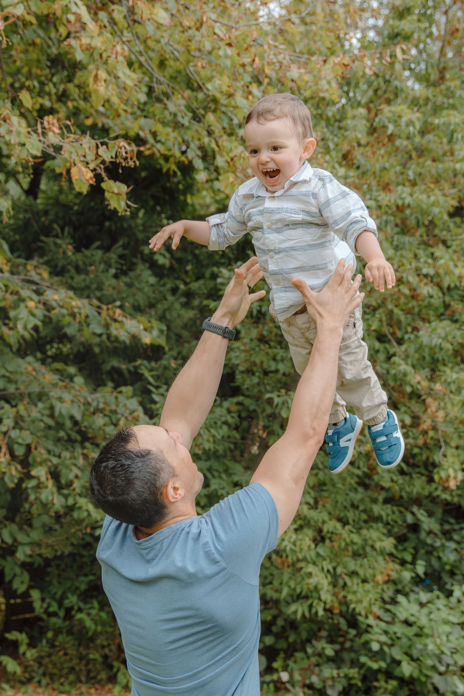 Joyful playtime as a child is lifted high into the air against a backdrop of green trees.