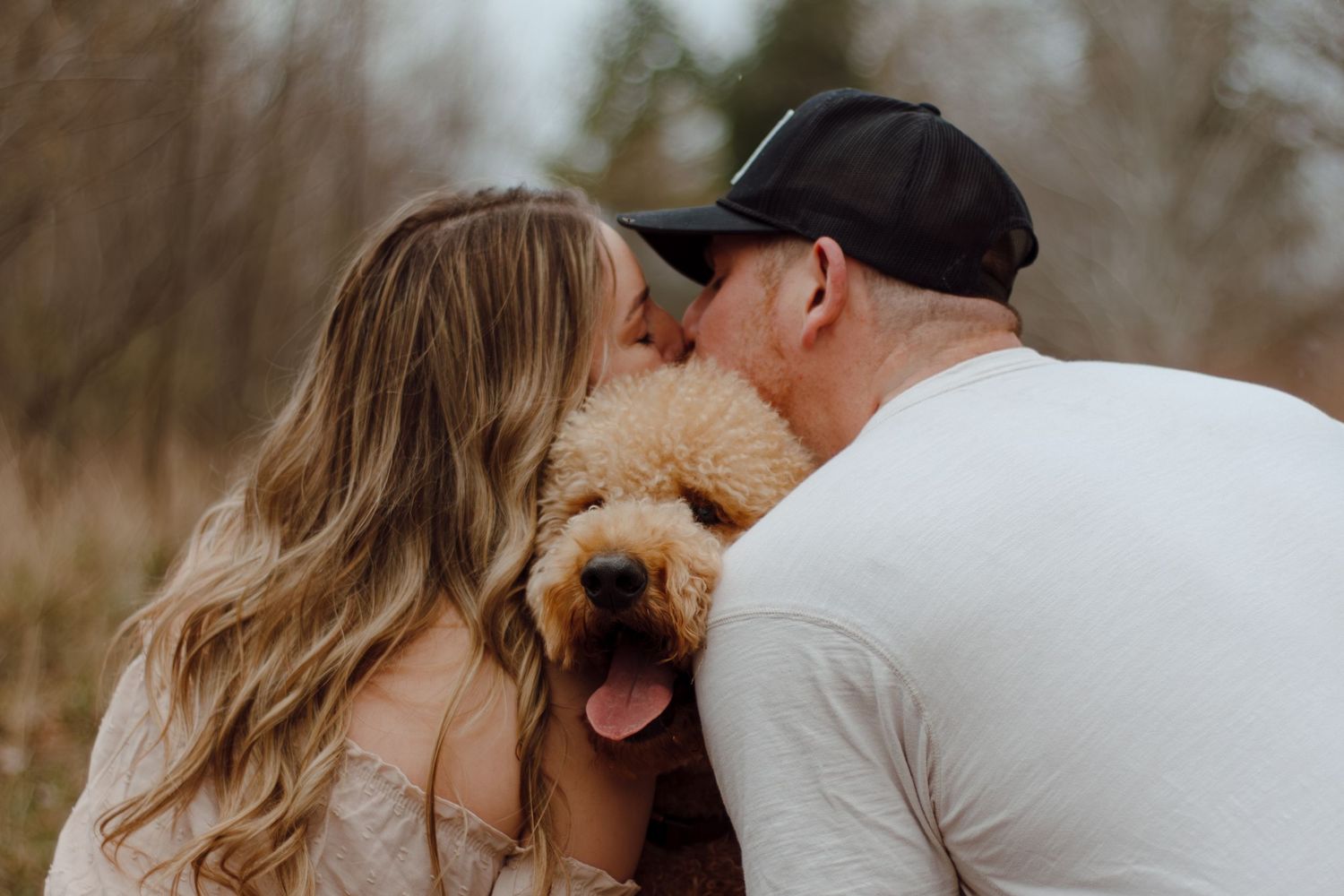 A loving couple kissing a fluffy golden doodle puppy outdoors in a romantic engagement photo session.