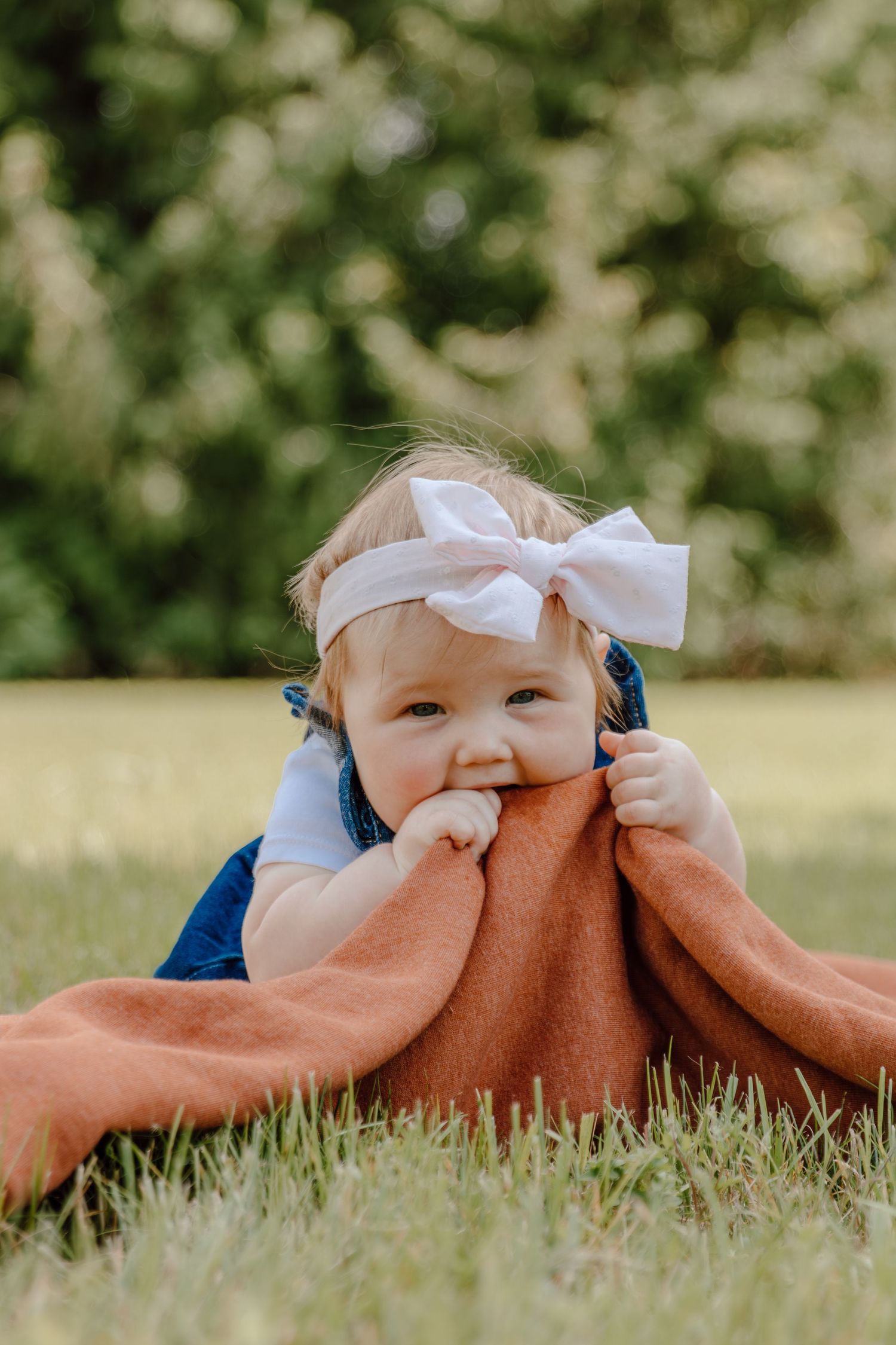Baby in blue overalls and white bow playing with orange blanket on grass.