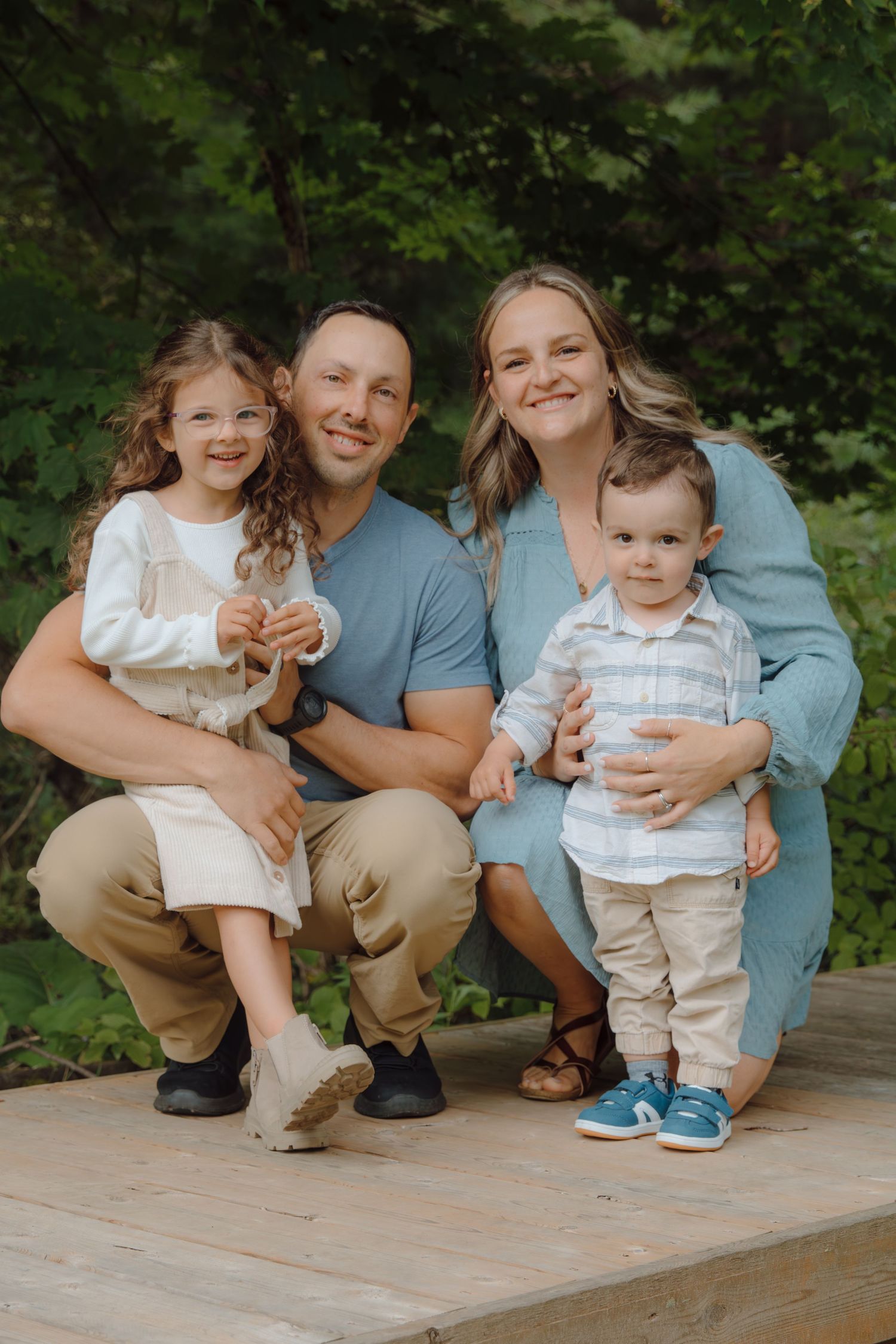 A family of four poses together on a wooden deck surrounded by lush green foliage.