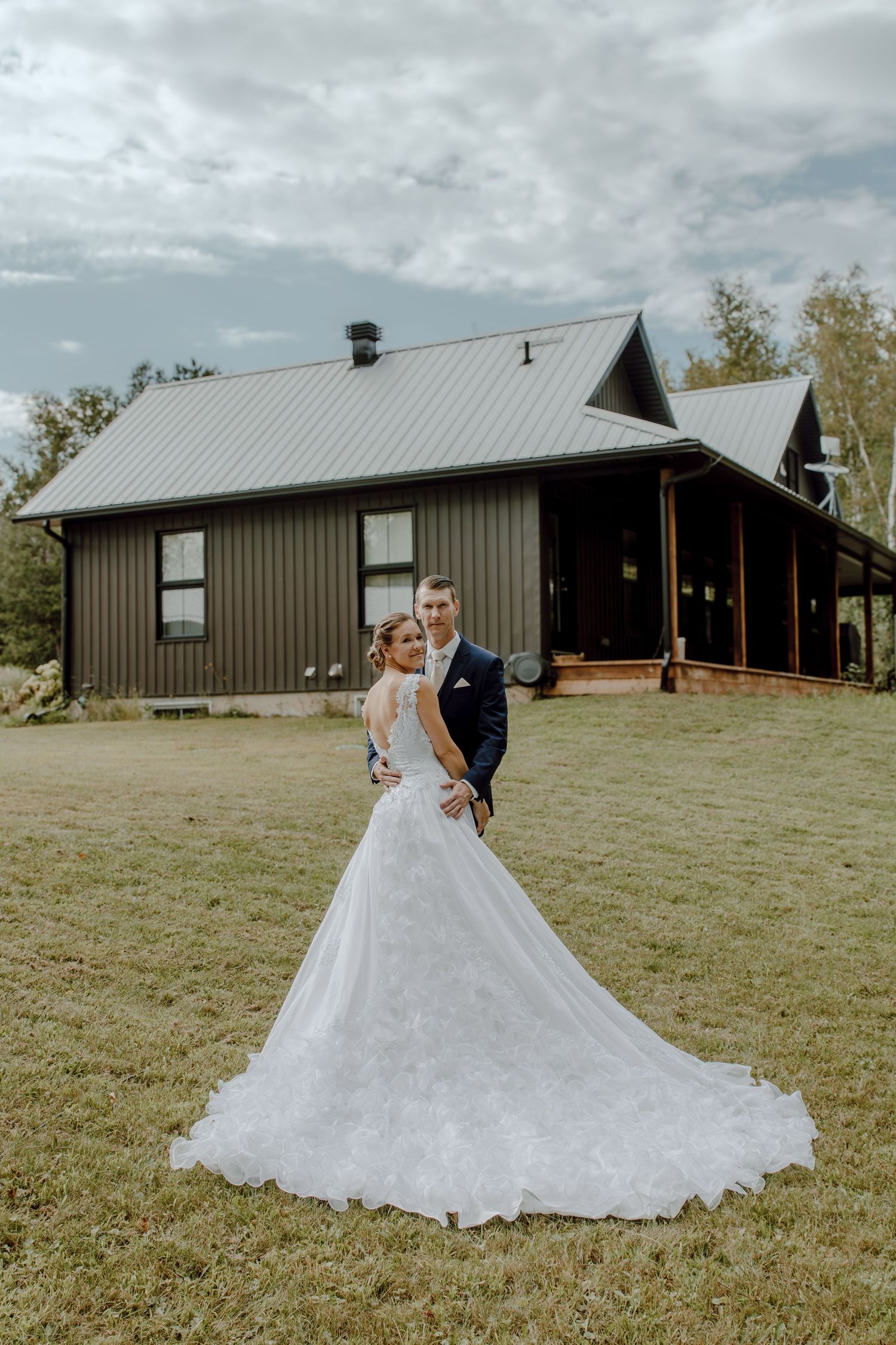 A couple in wedding attire dances together on a grassy field in front of a modern barn-style building.
