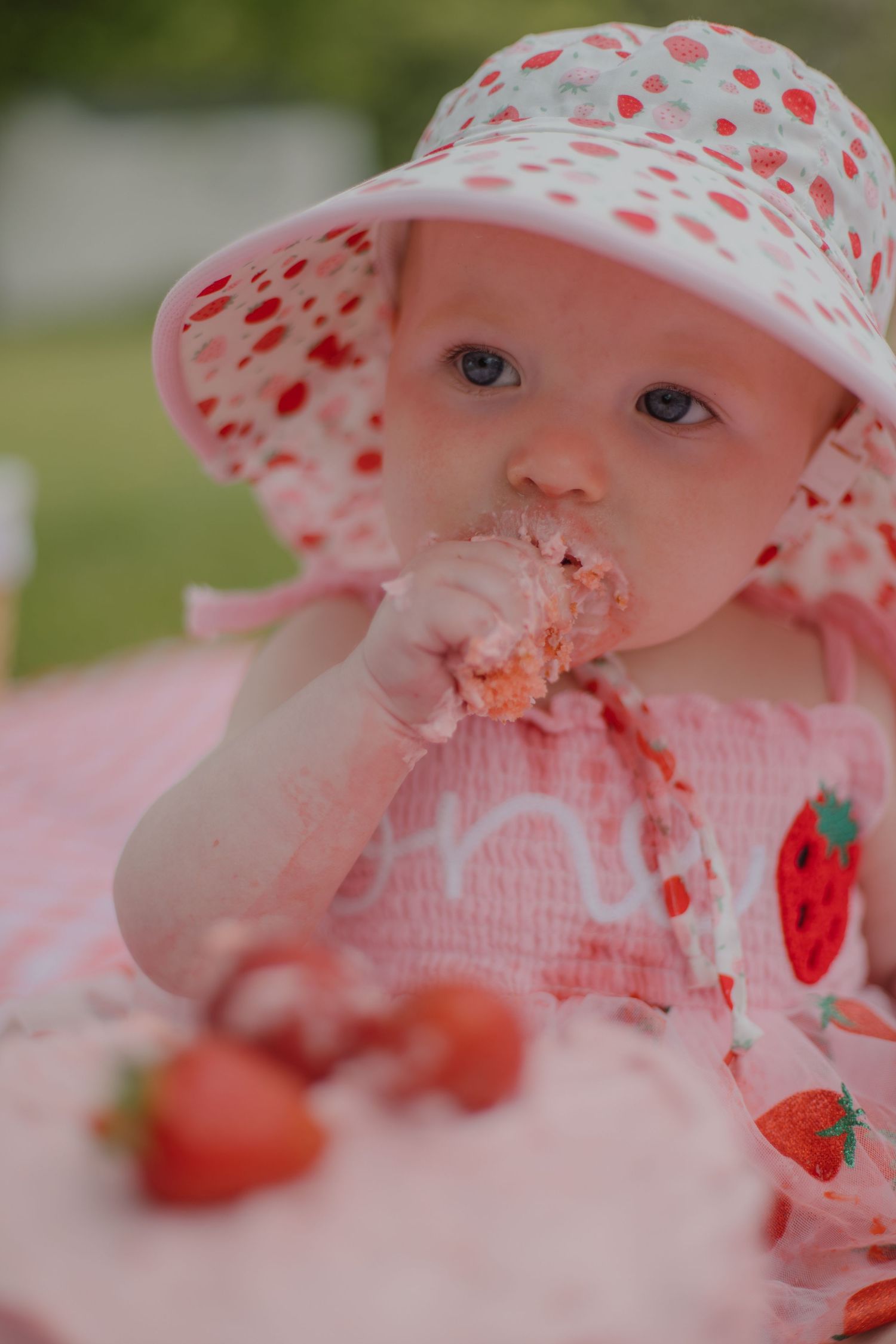 Baby girl in strawberry hat and dress eating cake at a first birthday party outdoors.