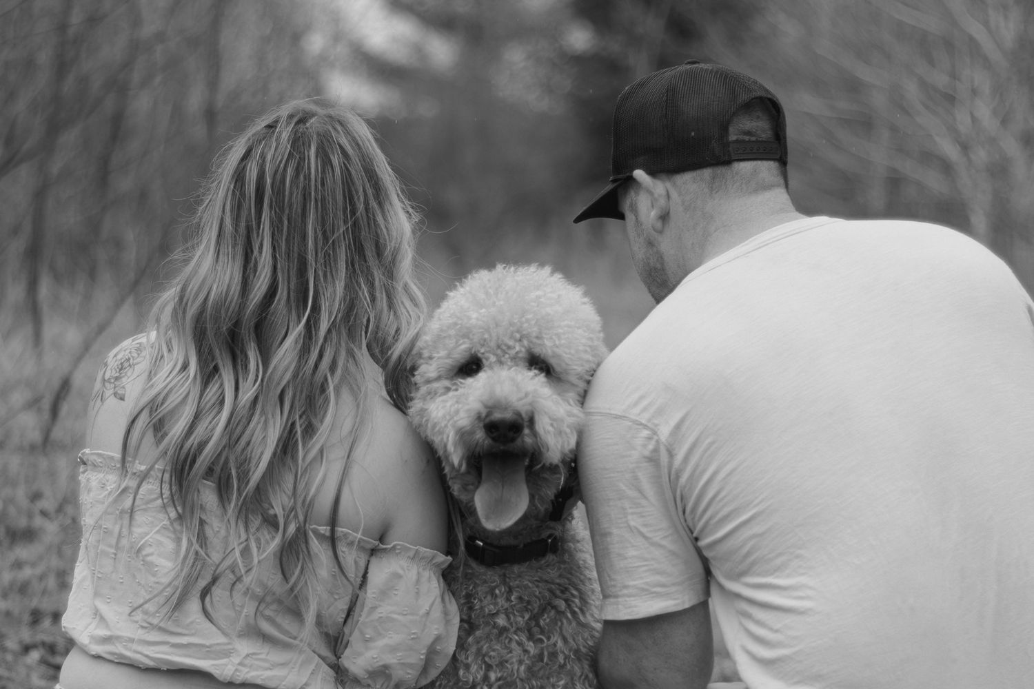 Black and white photo of a couple sitting with their fluffy goldendoodle dog outdoors in a wooded setting.