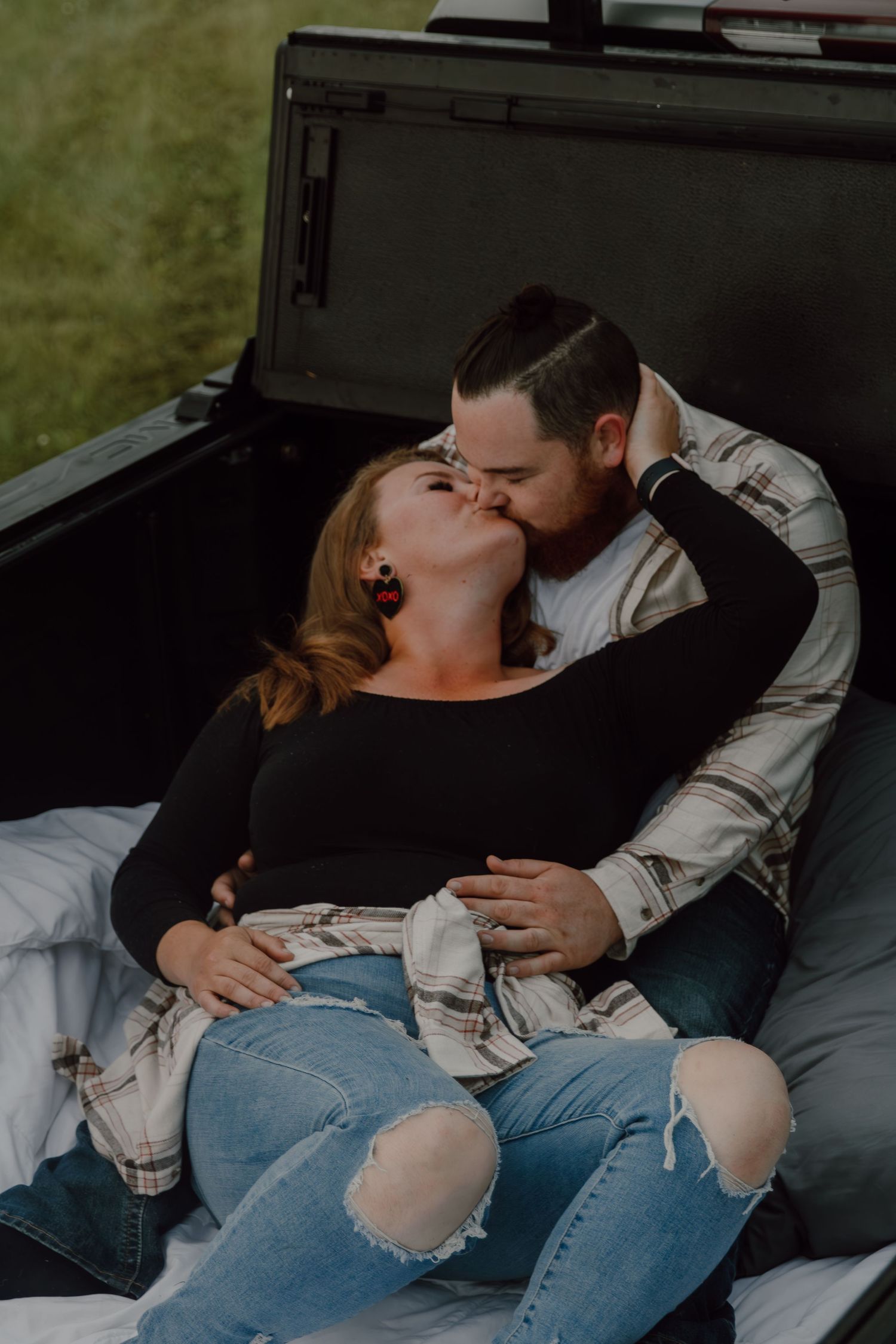 A couple sharing a romantic kiss while relaxing in the back of a truck bed, woman in ripped jeans and black top.