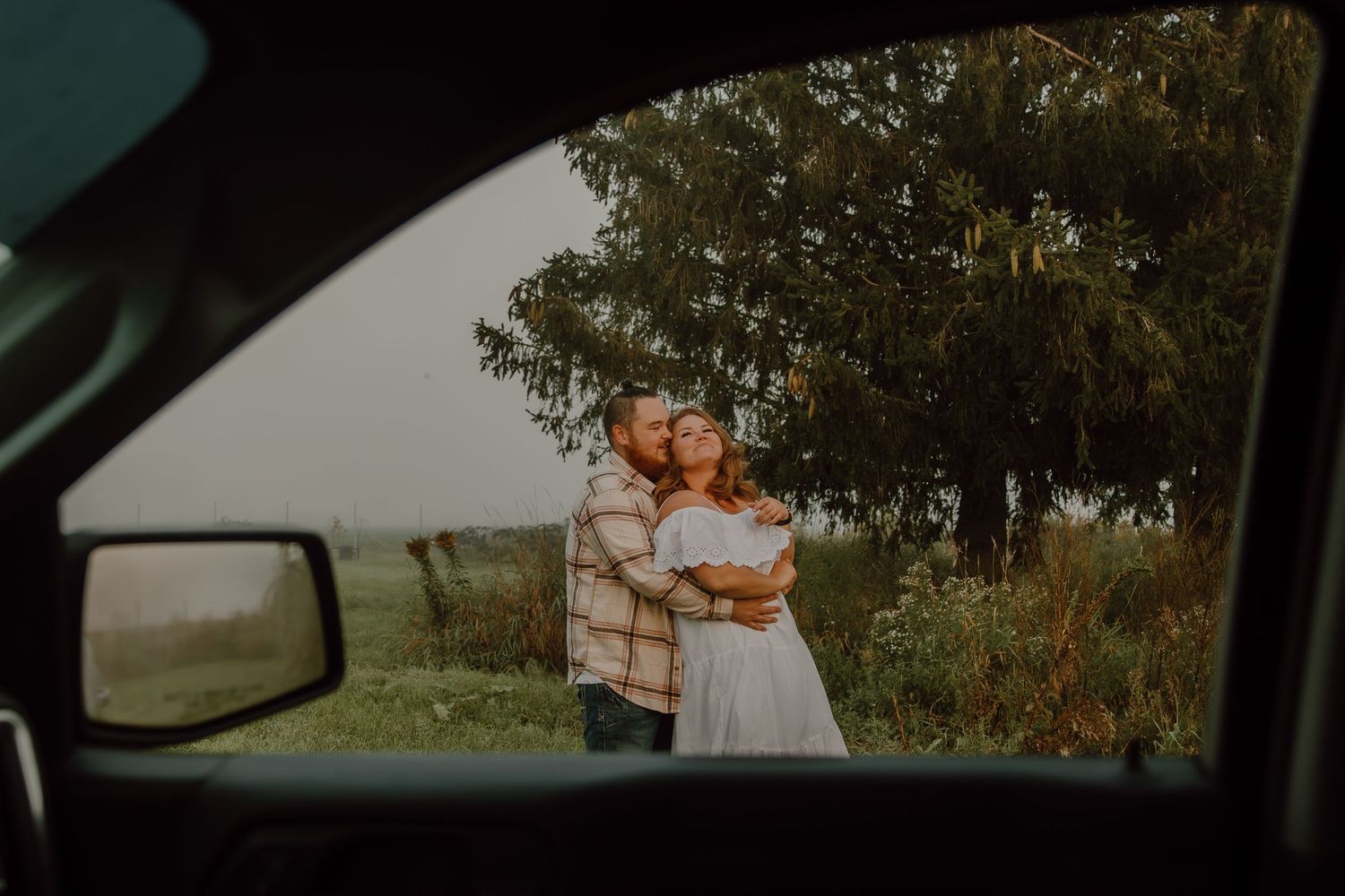 Couple embracing outdoors near trees, photographed through a car window with side mirror visible in foreground.