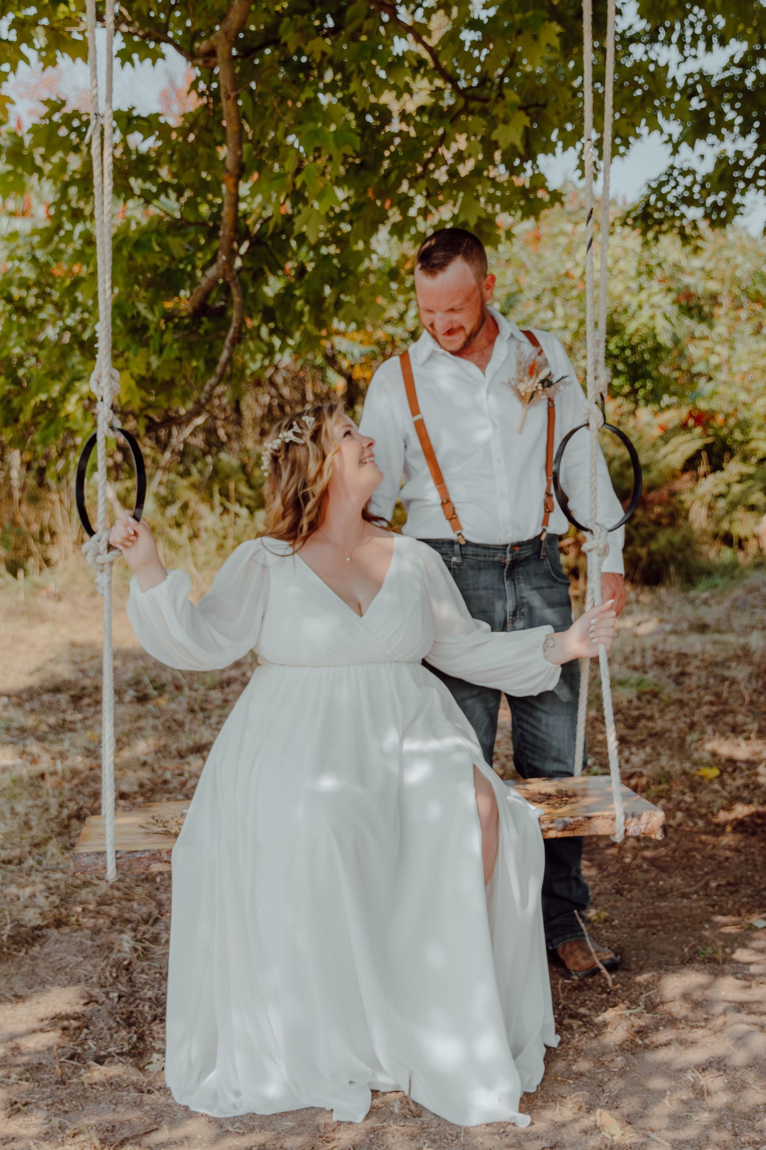 A couple shares a romantic moment on a wooden swing surrounded by trees in warm sunlight.
