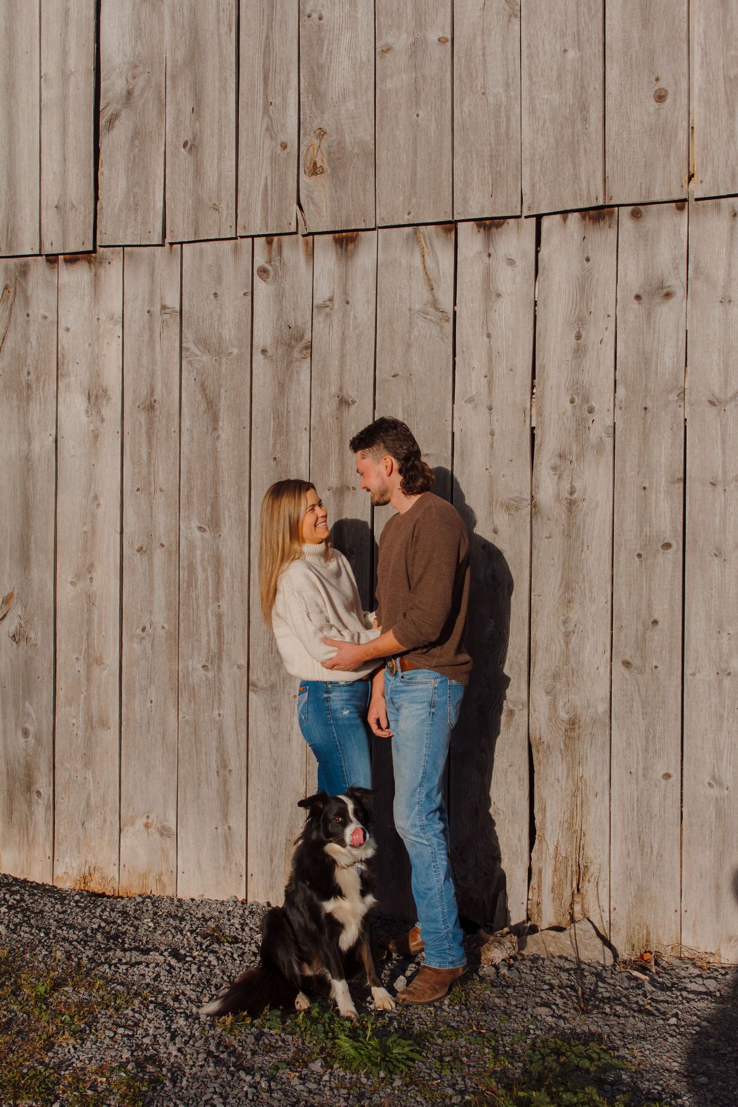 Two people embrace by a weathered barn wall while their dog watches.
