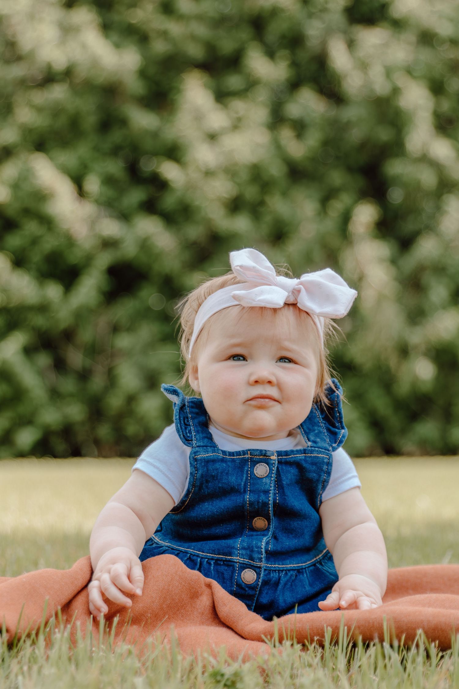 Baby in denim overalls and white bow sitting on orange blanket outdoors.