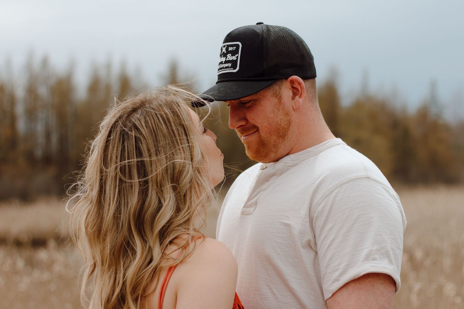 A couple shares a romantic moment in a golden field, the man wearing a black cap and white t-shirt leaning toward a blonde woman.
