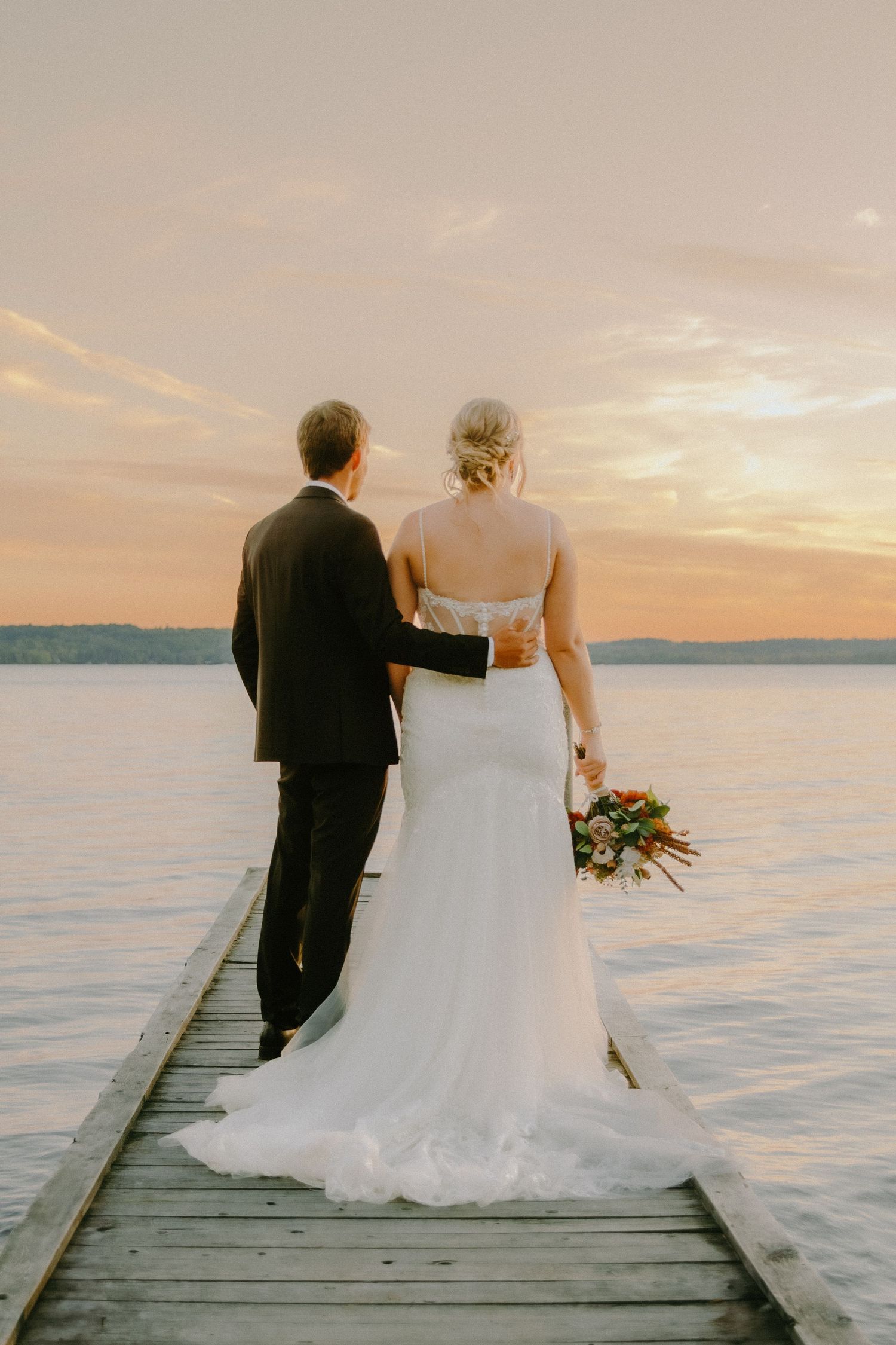 Newlyweds stand together on a wooden pier overlooking a serene lake at sunset with warm golden light.