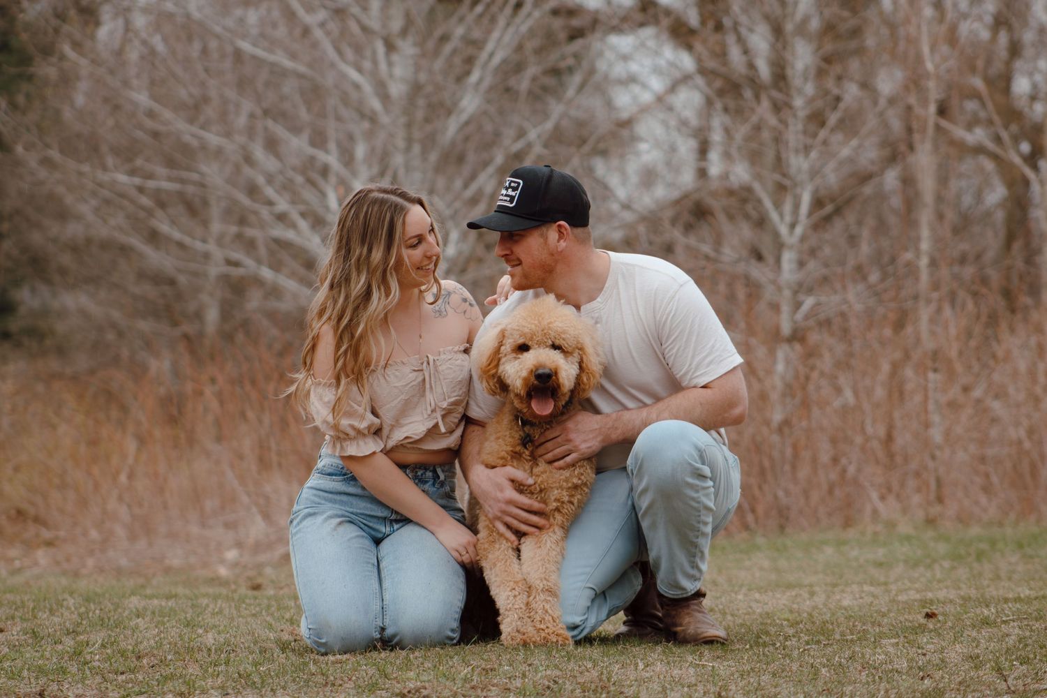 Couple posing outdoors with golden doodle dog in a field with bare trees in background.