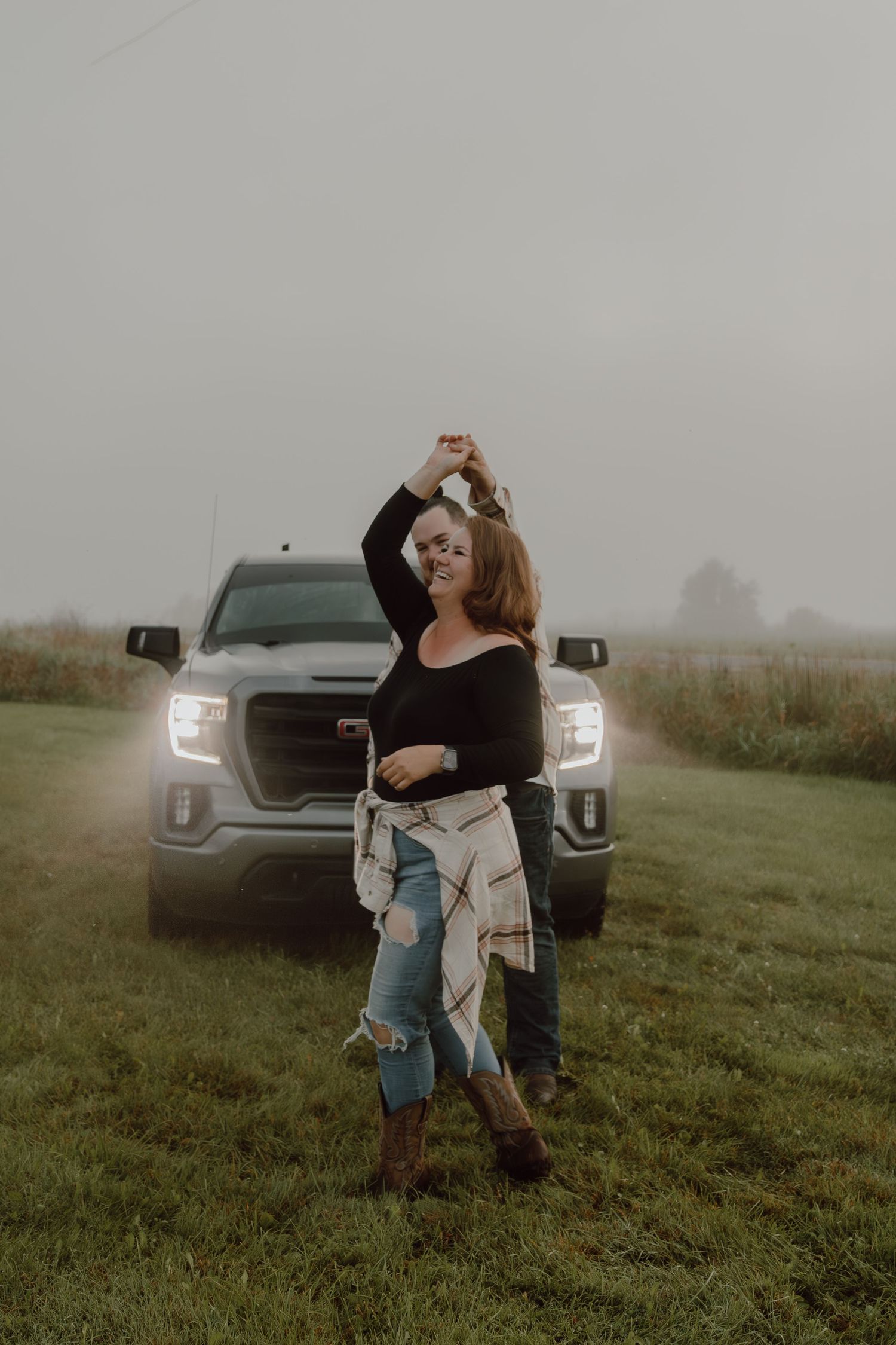 Woman dancing in a field beside a GMC truck on a foggy day, wearing ripped jeans and cowboy boots.