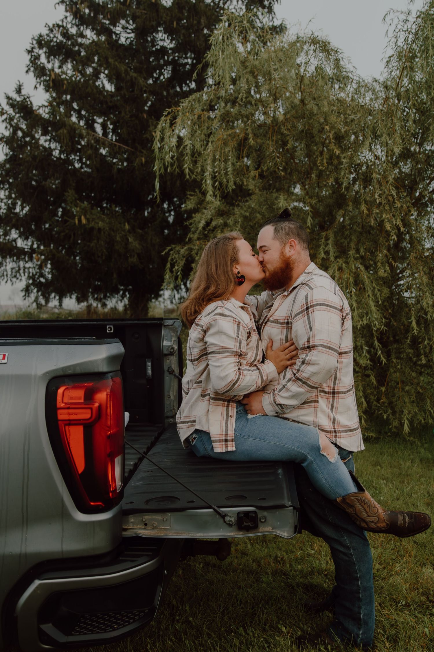 Couple kissing on the tailgate of a silver GMC truck, wearing matching plaid shirts and jeans, surrounded by lush green trees.