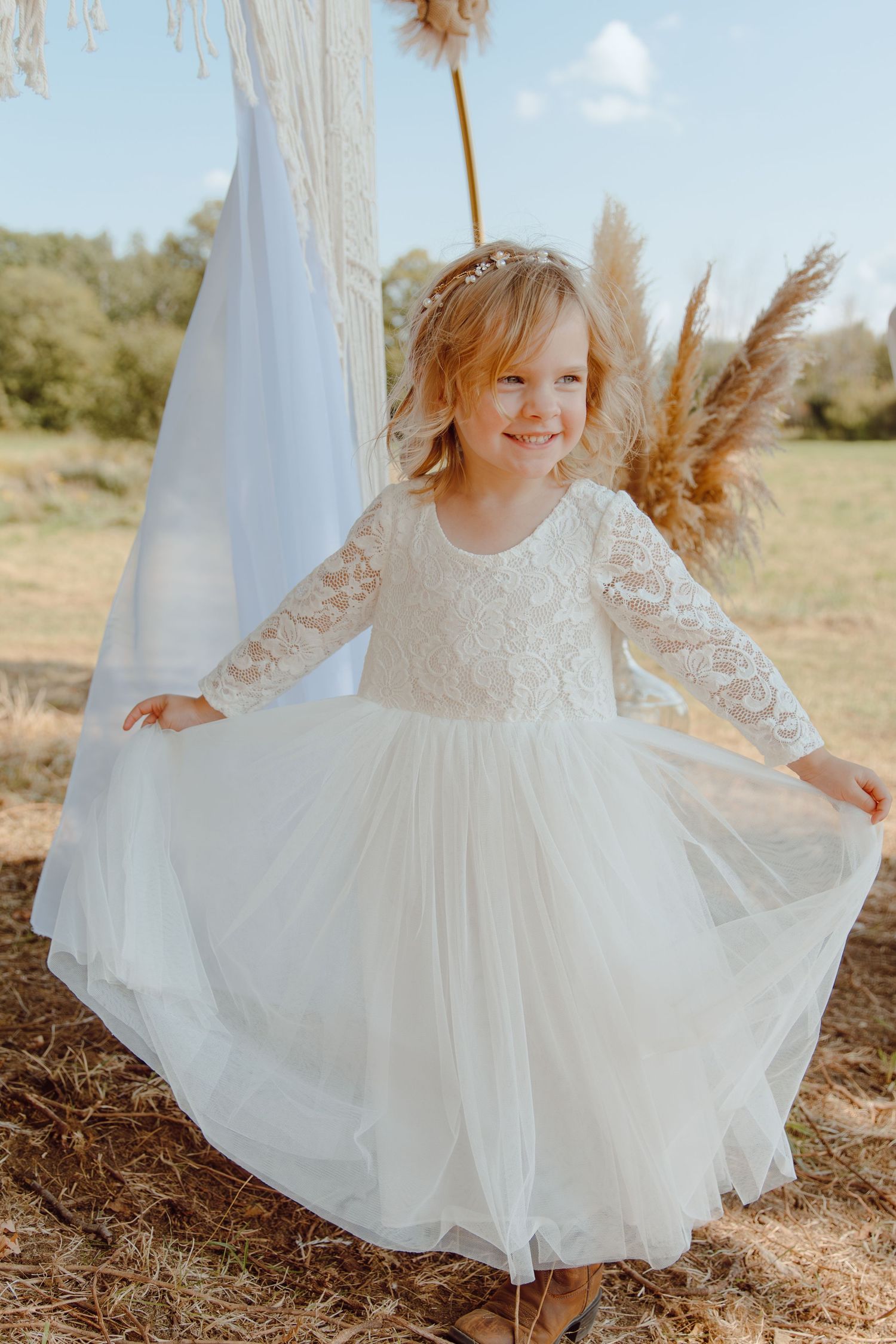 A young child in a white lace dress twirls joyfully outdoors in natural sunlight.