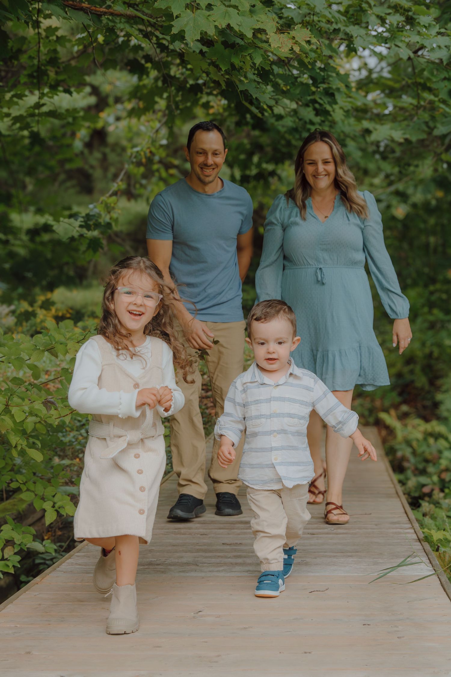 A family runs and plays together on a wooden boardwalk surrounded by green foliage.