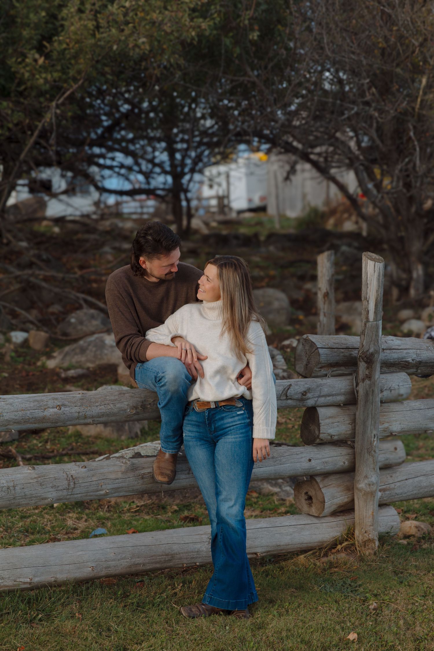 A romantic autumn scene with a couple leaning against a wooden fence as trees sway in the background.