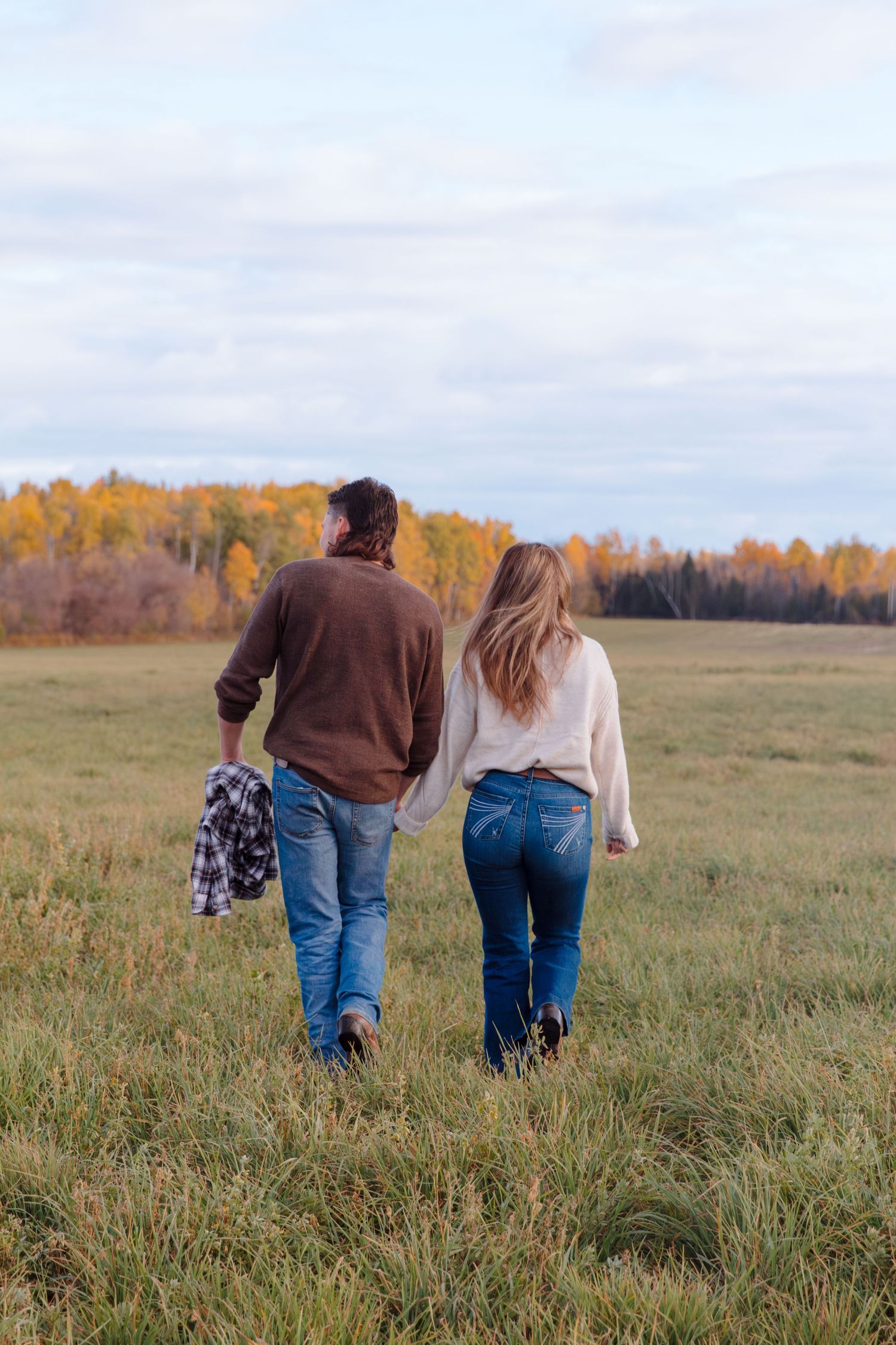 A couple strolls through a scenic autumn field with golden trees on the horizon.