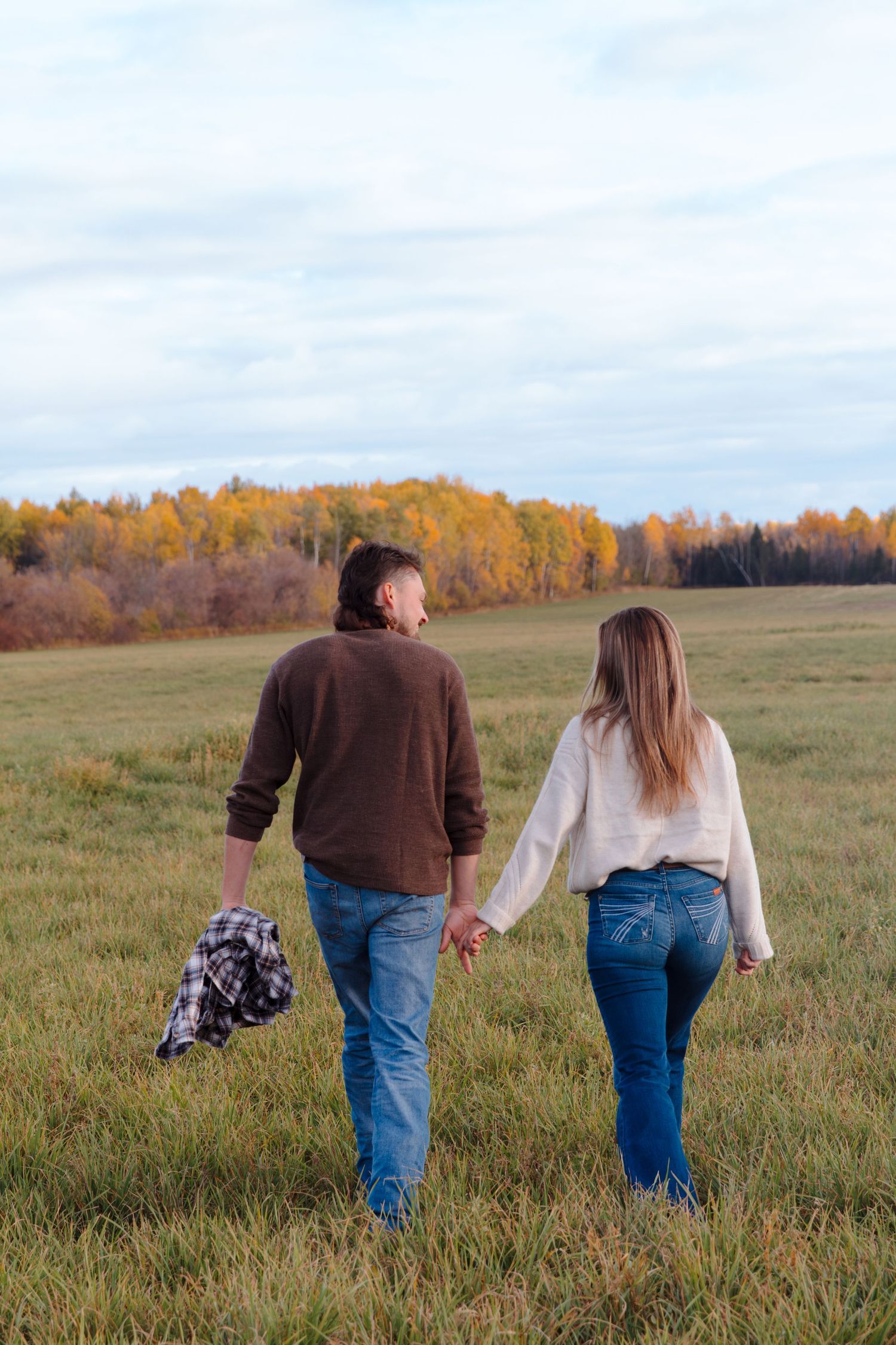 A couple walks hand in hand through a grassy field with golden autumn trees in the distance.