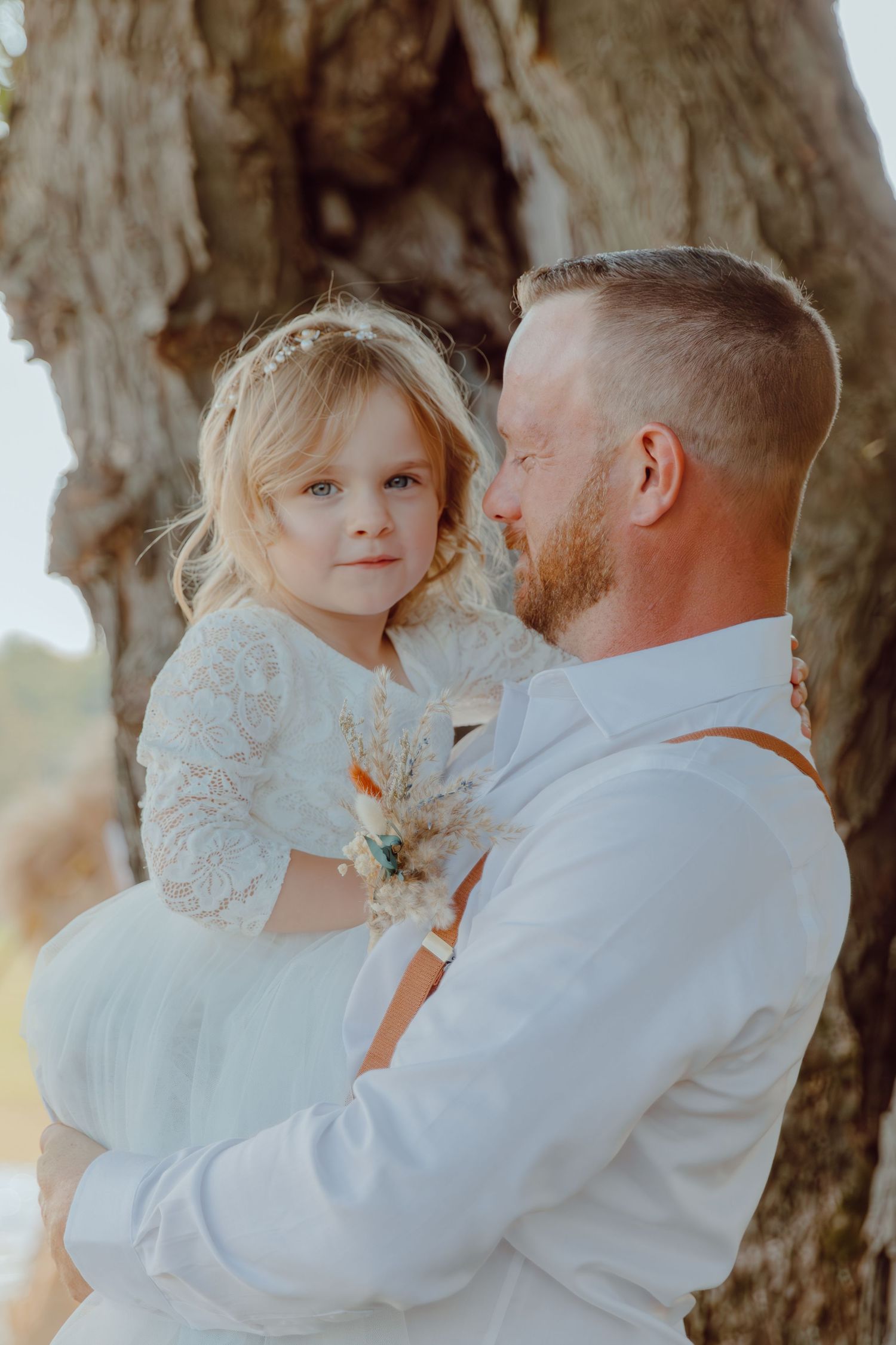 A tender family moment captured in soft lighting under a tree, with white clothing creating an ethereal atmosphere.