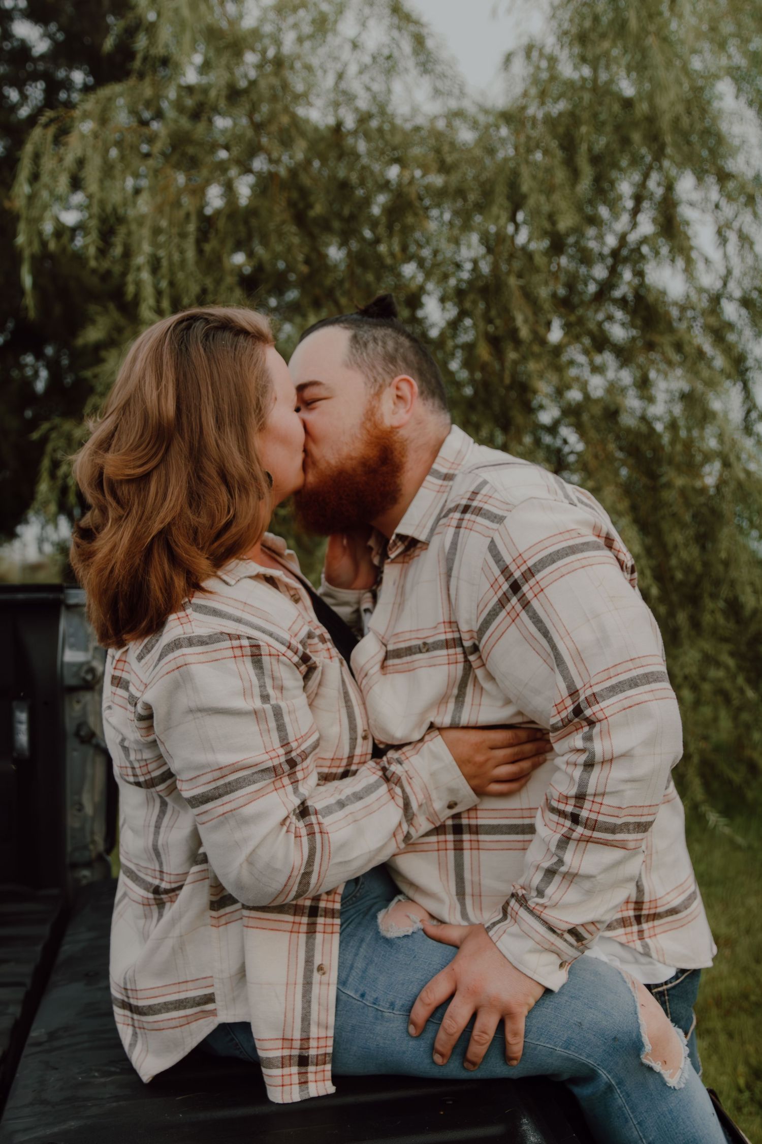 A couple sharing a romantic kiss outdoors, both wearing matching plaid flannel shirts, with a lush green willow tree in the background.