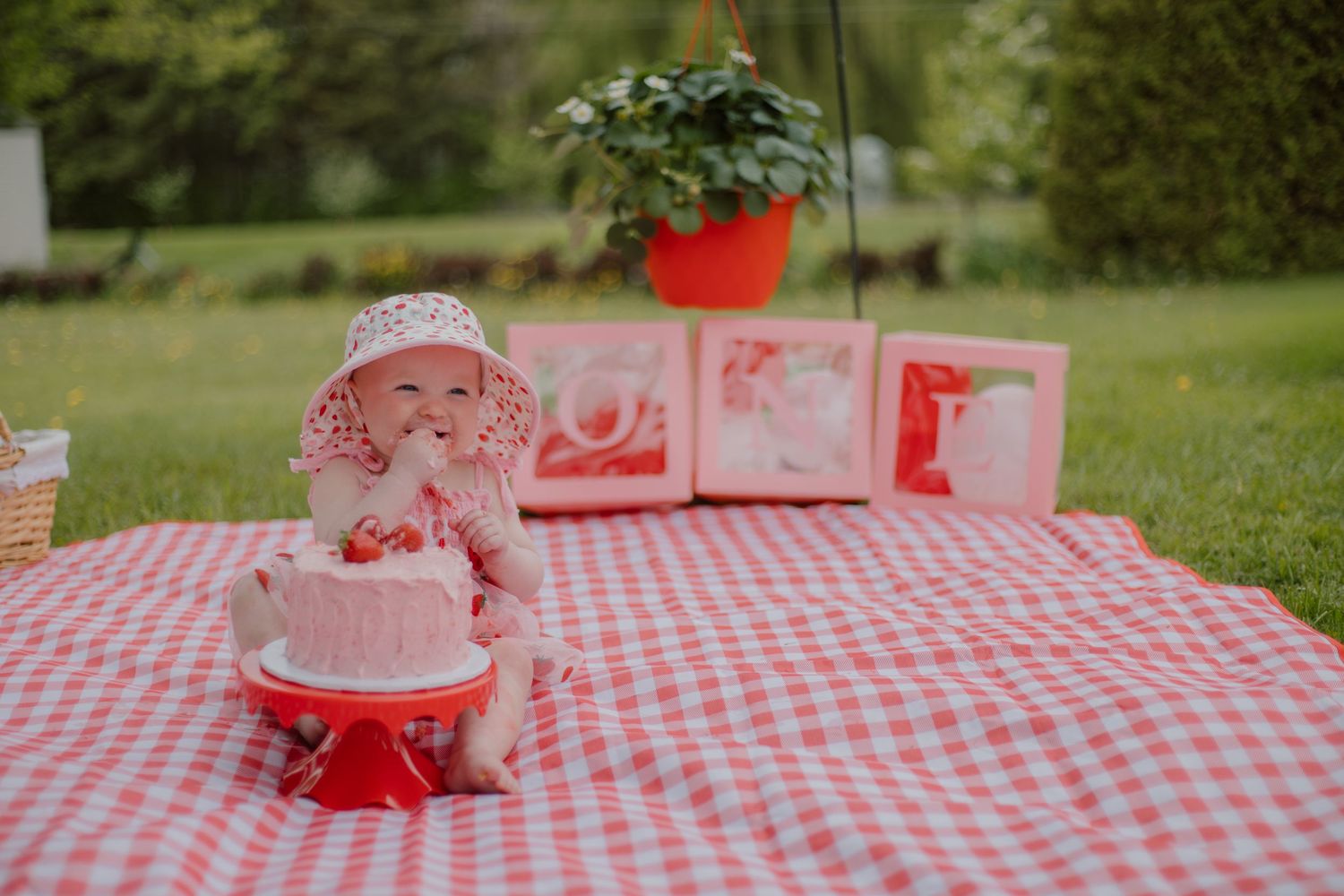 Baby girl in floral hat enjoys first birthday cake smash on red gingham blanket outdoors with strawberry decorations.