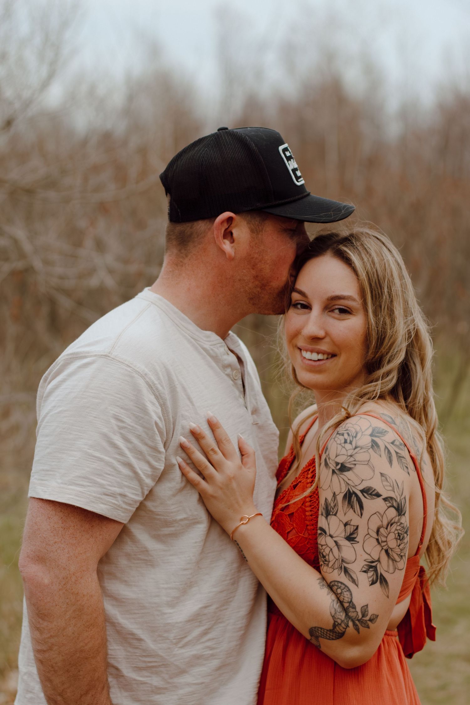 A couple embraces outdoors, woman in orange dress with floral arm tattoo smiles as man in black cap kisses her temple.