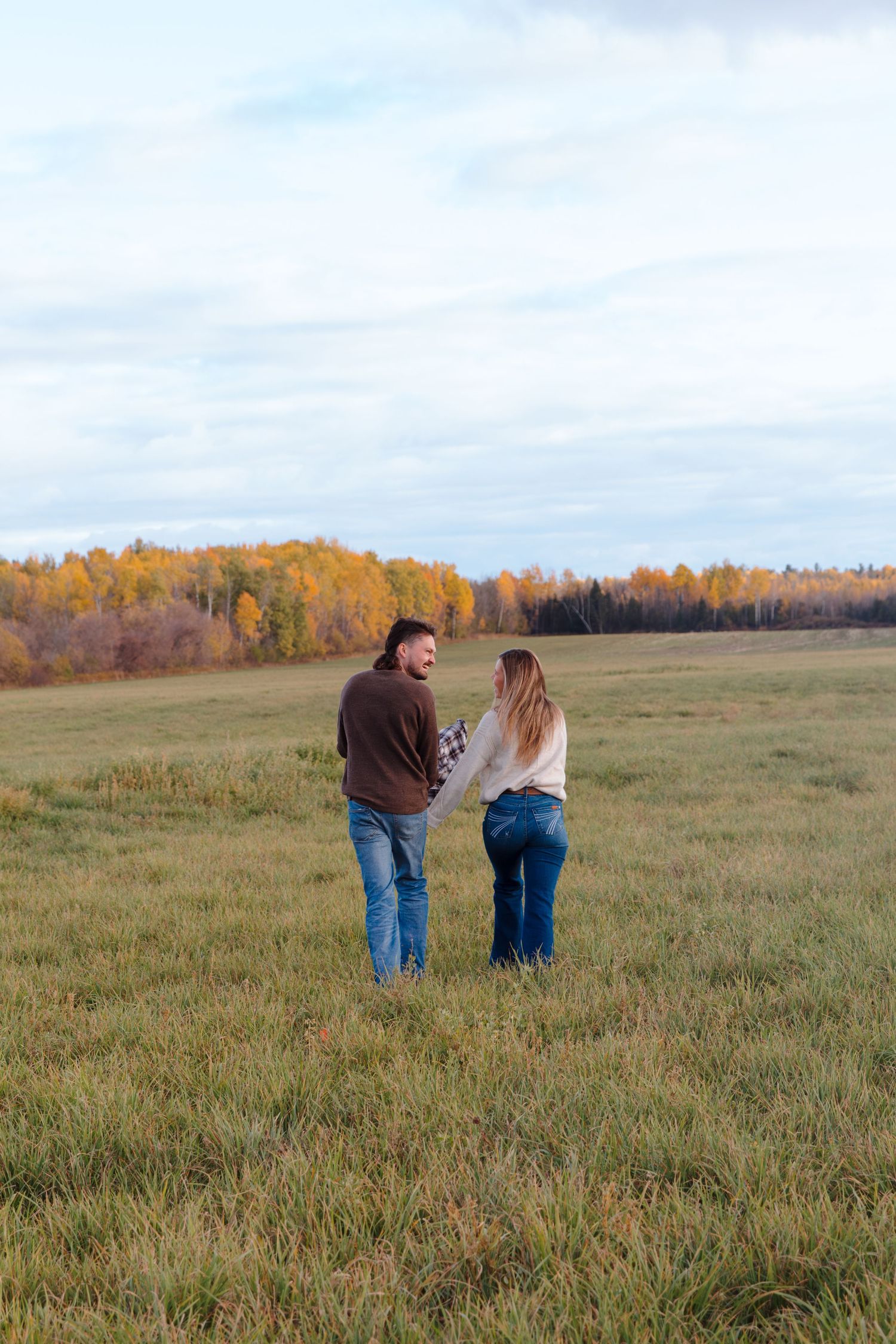 Two people walk together across a grassy field with autumn trees in the distance.