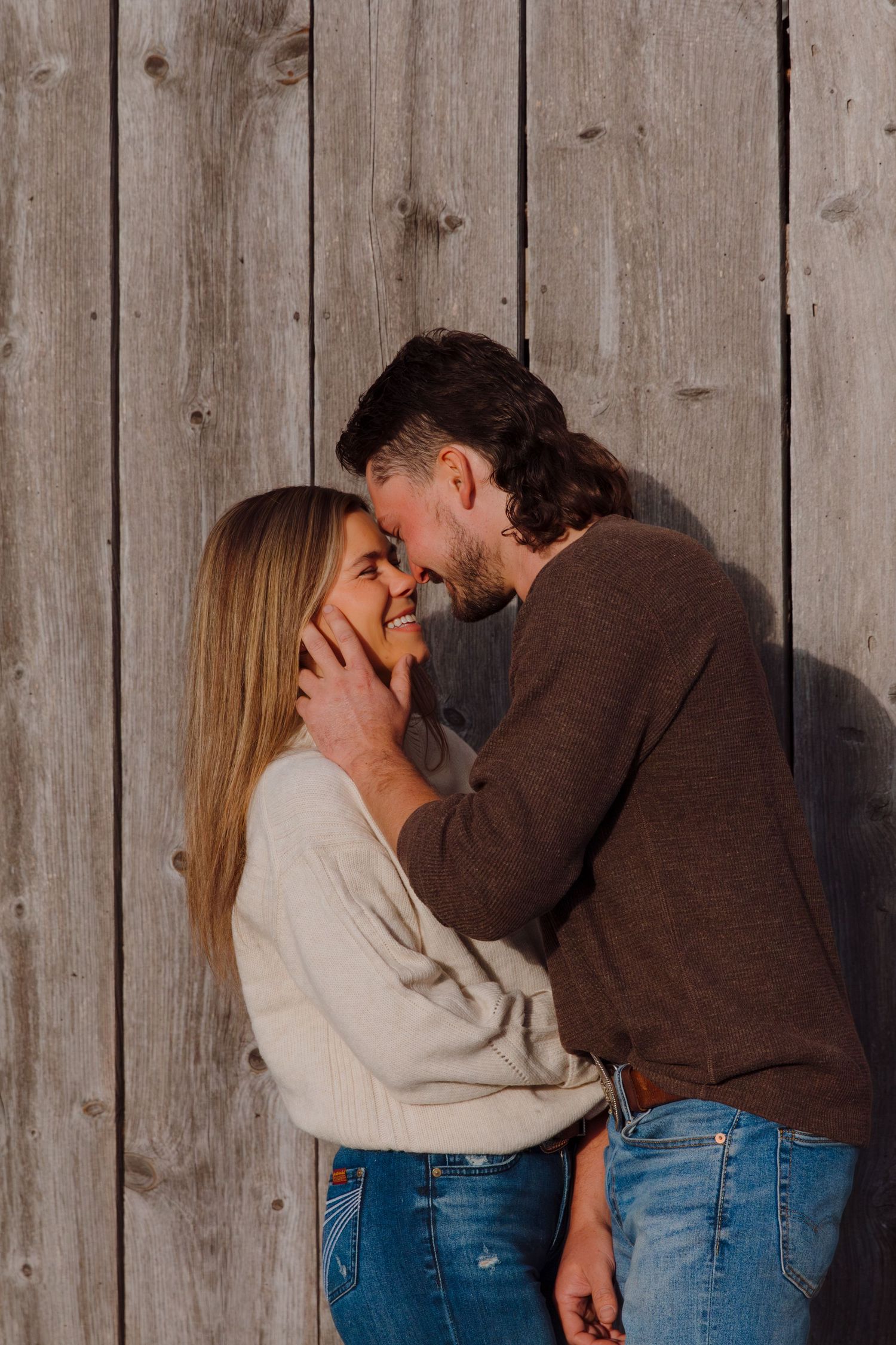 Two people share laughter and embrace against a wooden barn wall.