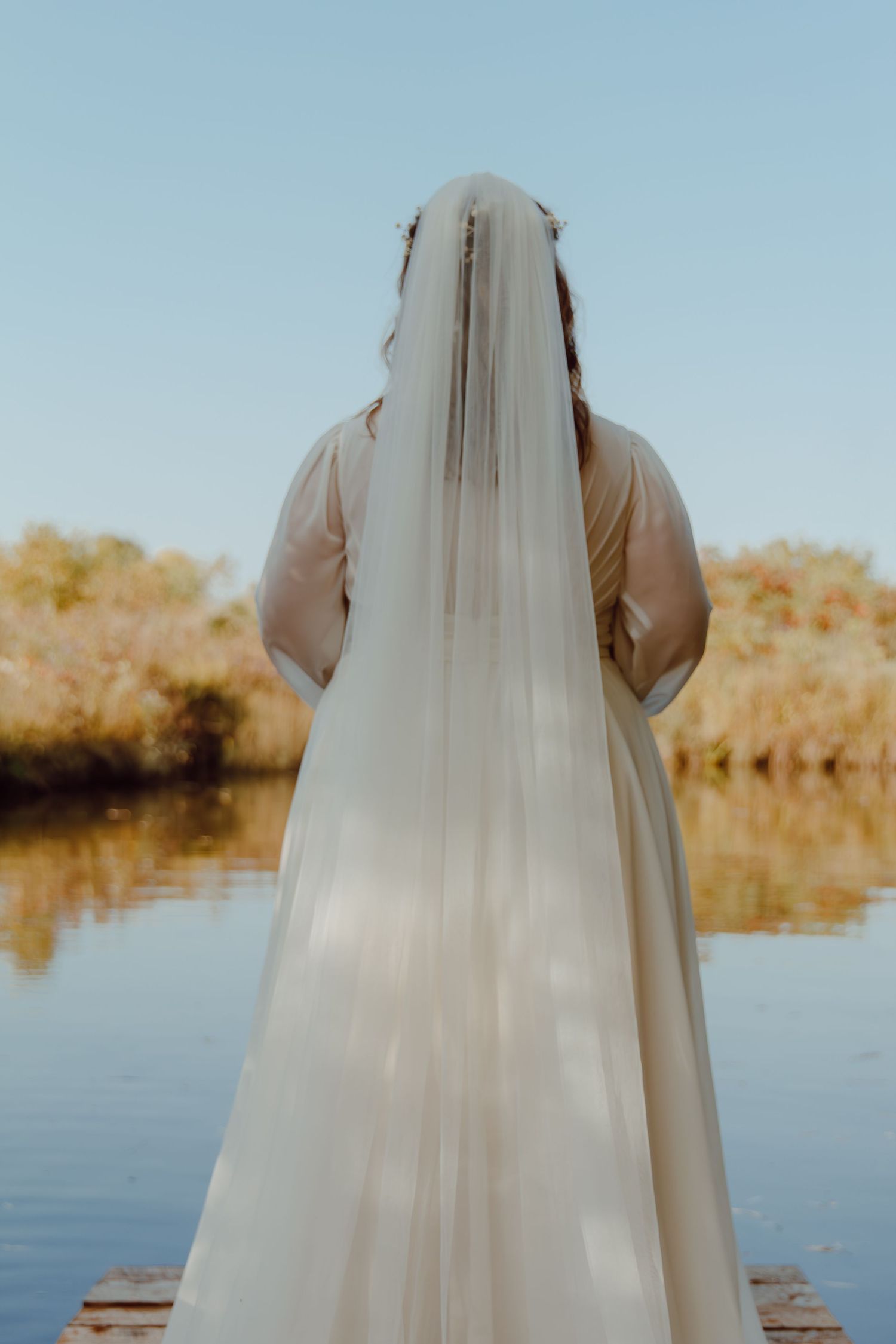 A bride in a flowing veil stands near a serene lake during sunset.