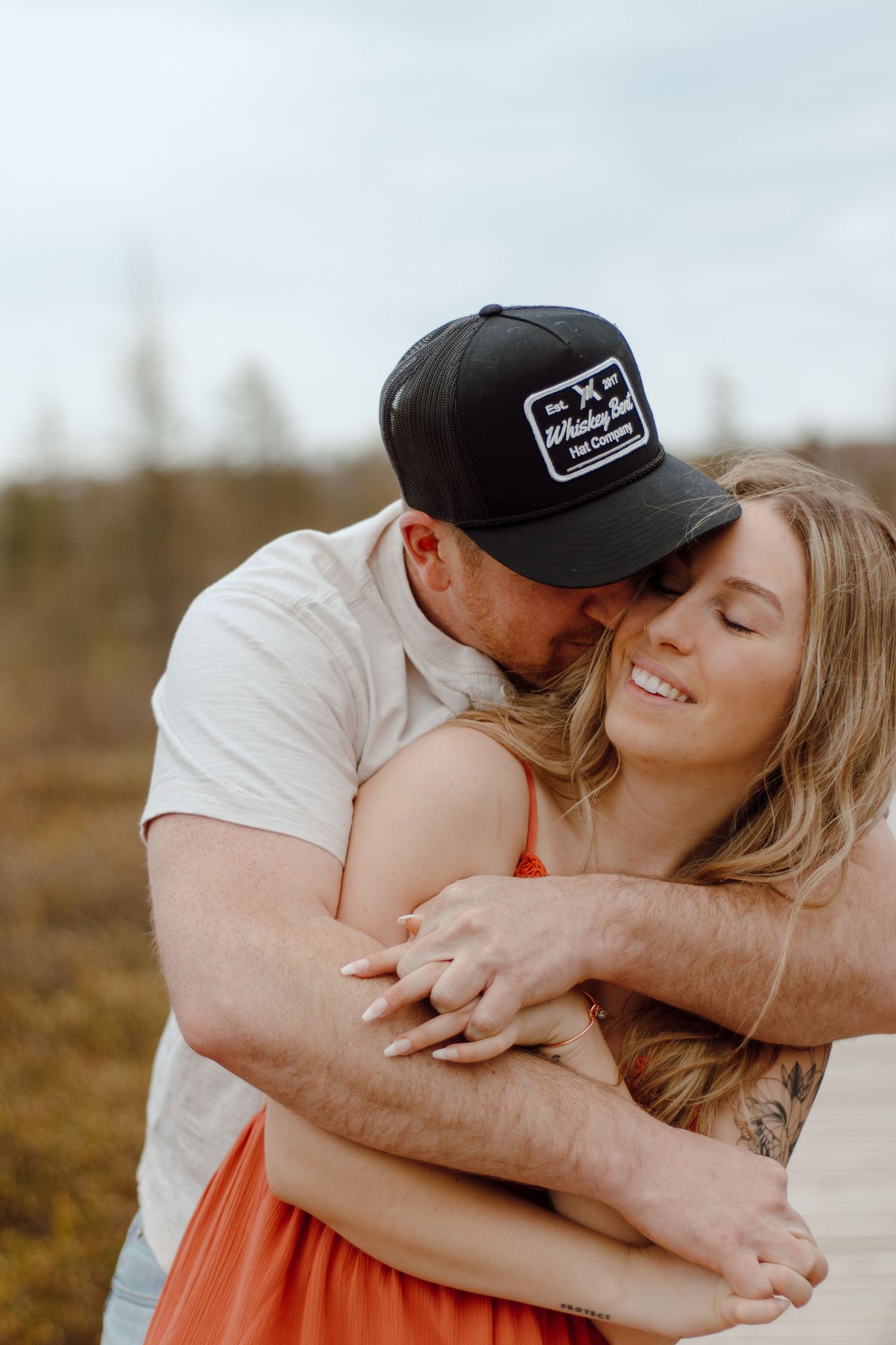 A couple shares a tender embrace outdoors, man in black cap kissing woman's cheek as she smiles, wearing an orange dress.