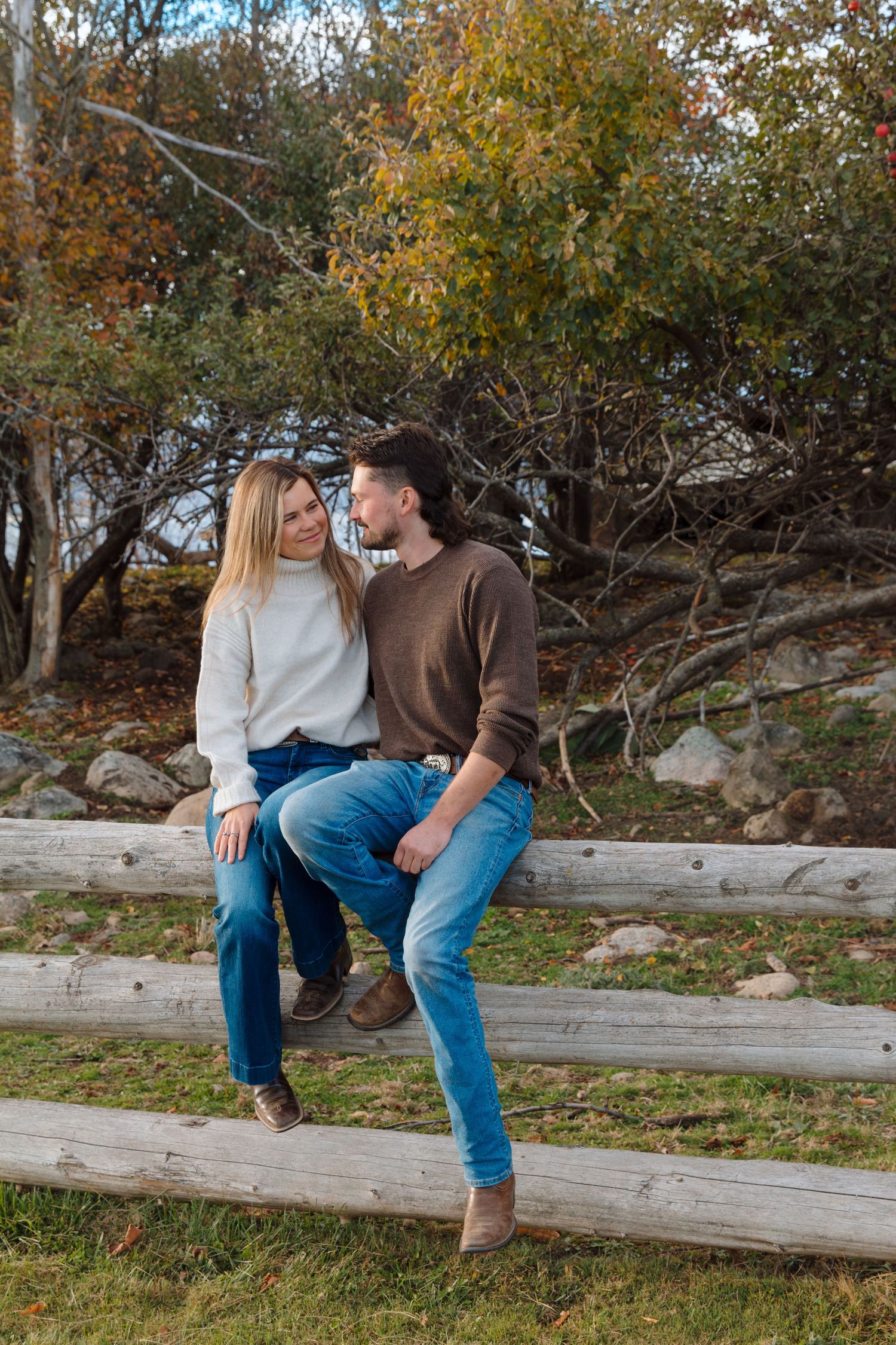A couple sits together on a rustic wooden fence surrounded by autumn trees and fallen leaves.