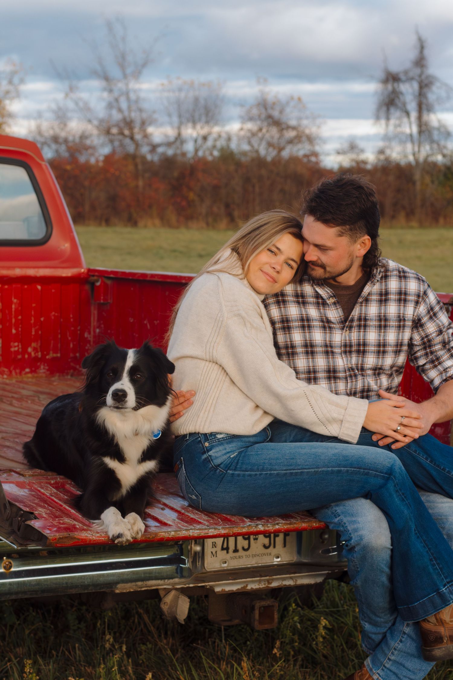 A couple and their Border Collie dog share tender moments while relaxing in the bed of a red pickup truck on an autumn day.