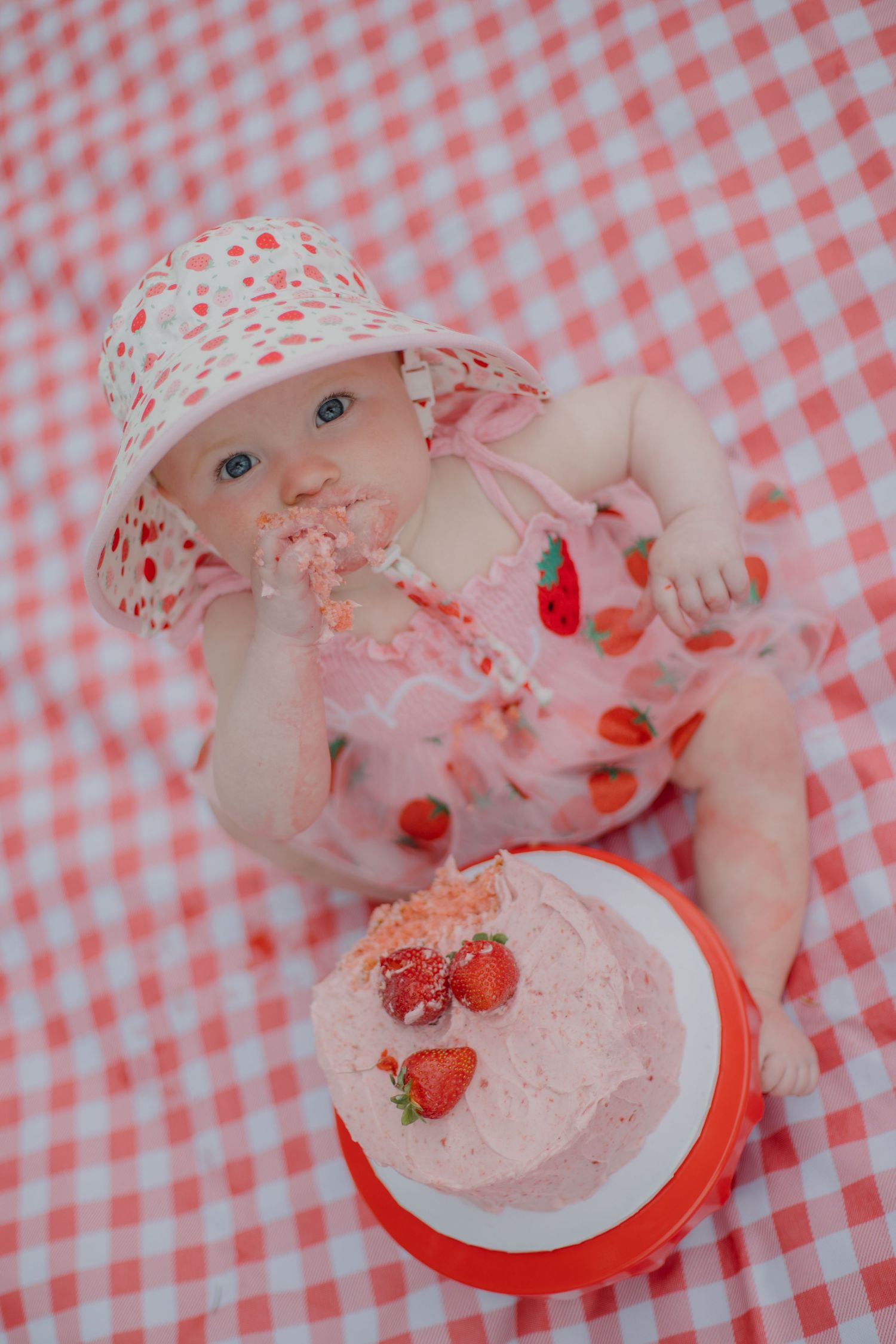 Baby girl in strawberry outfit smashes pink strawberry cake on red gingham blanket during first birthday smash cake photo.
