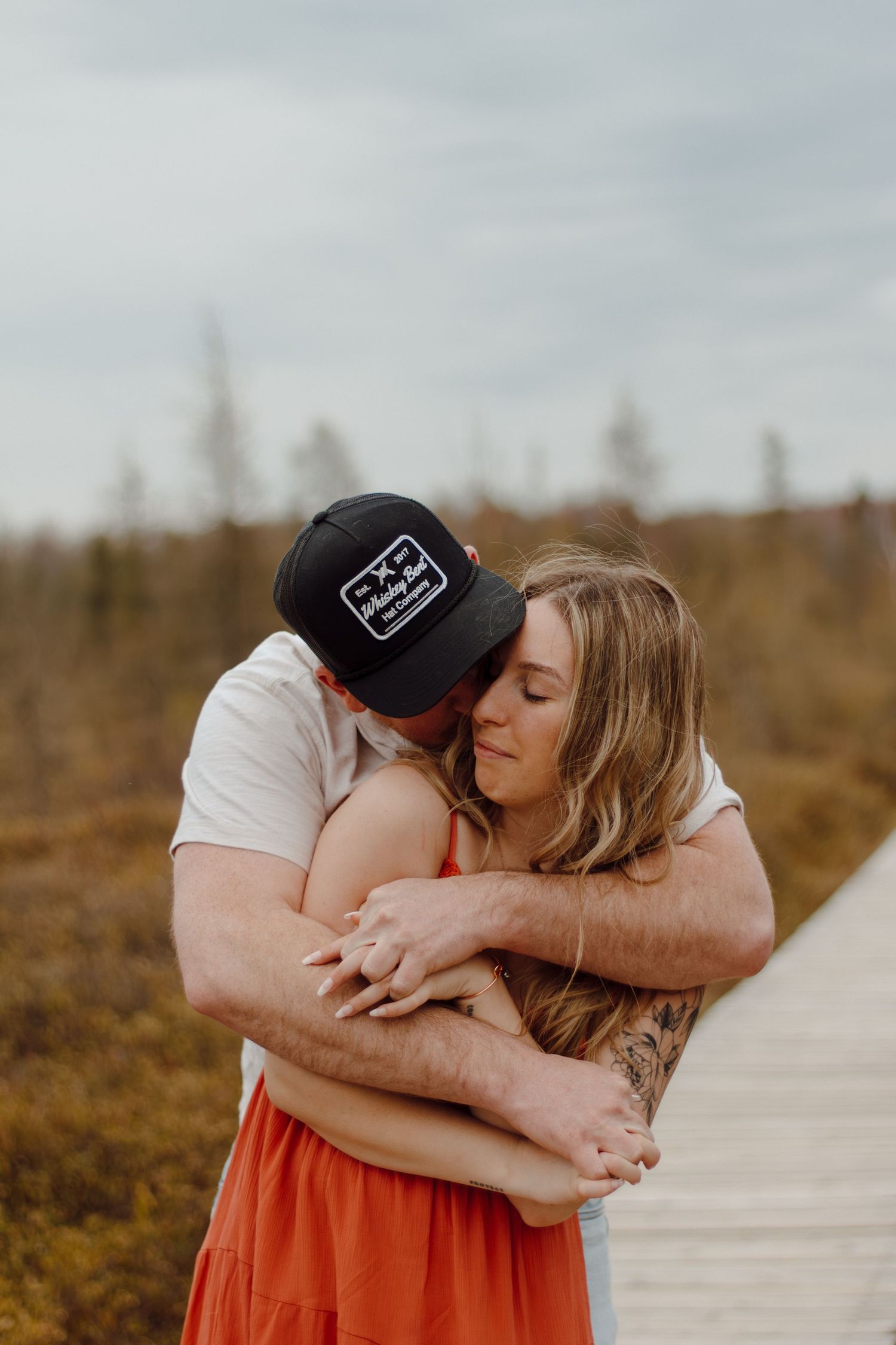 A couple shares a tender embrace on a wooden boardwalk surrounded by autumn fields, the man kissing her cheek.