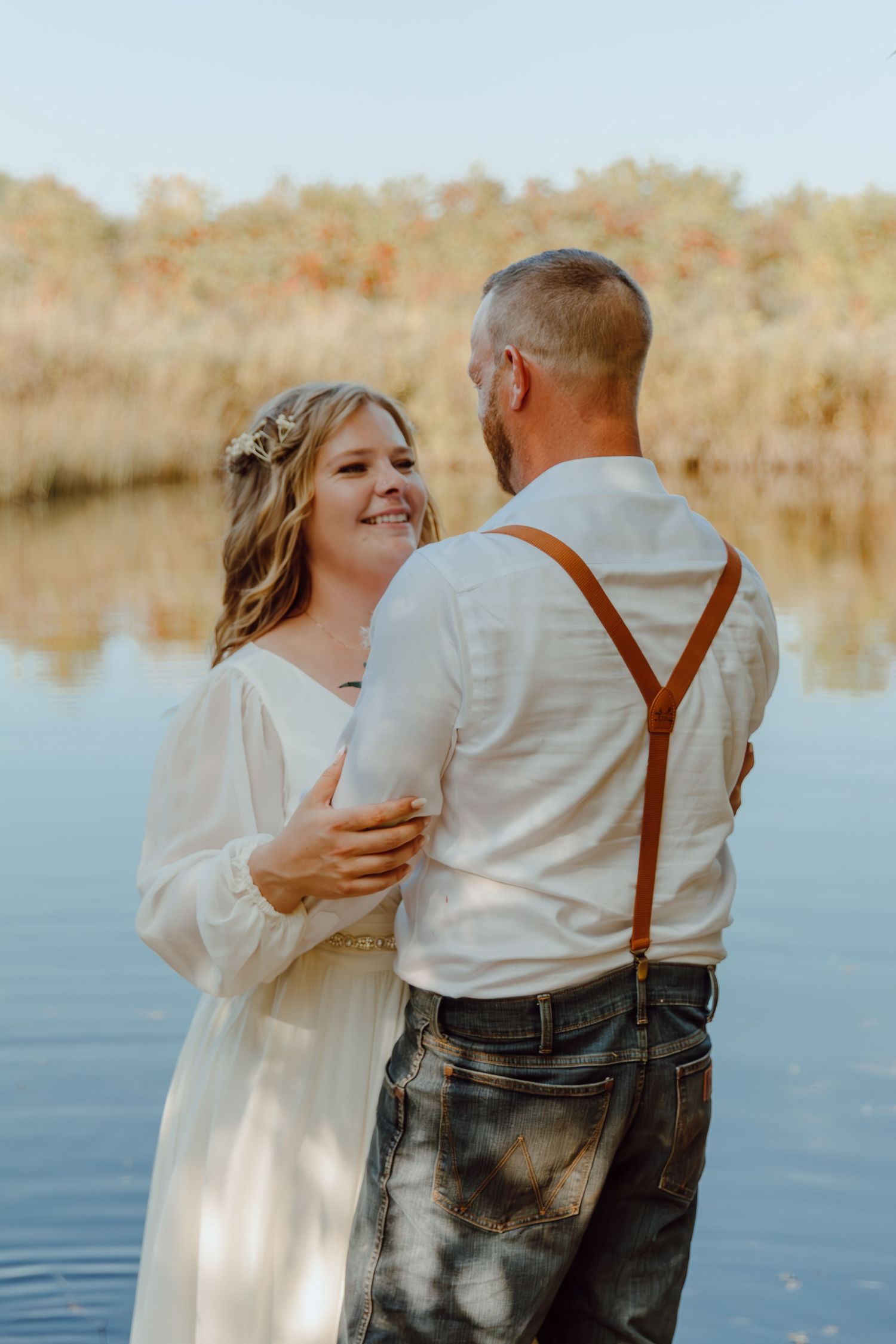 A romantic couple embraces by a serene lake during autumn, wearing white and brown vintage-inspired outfits.