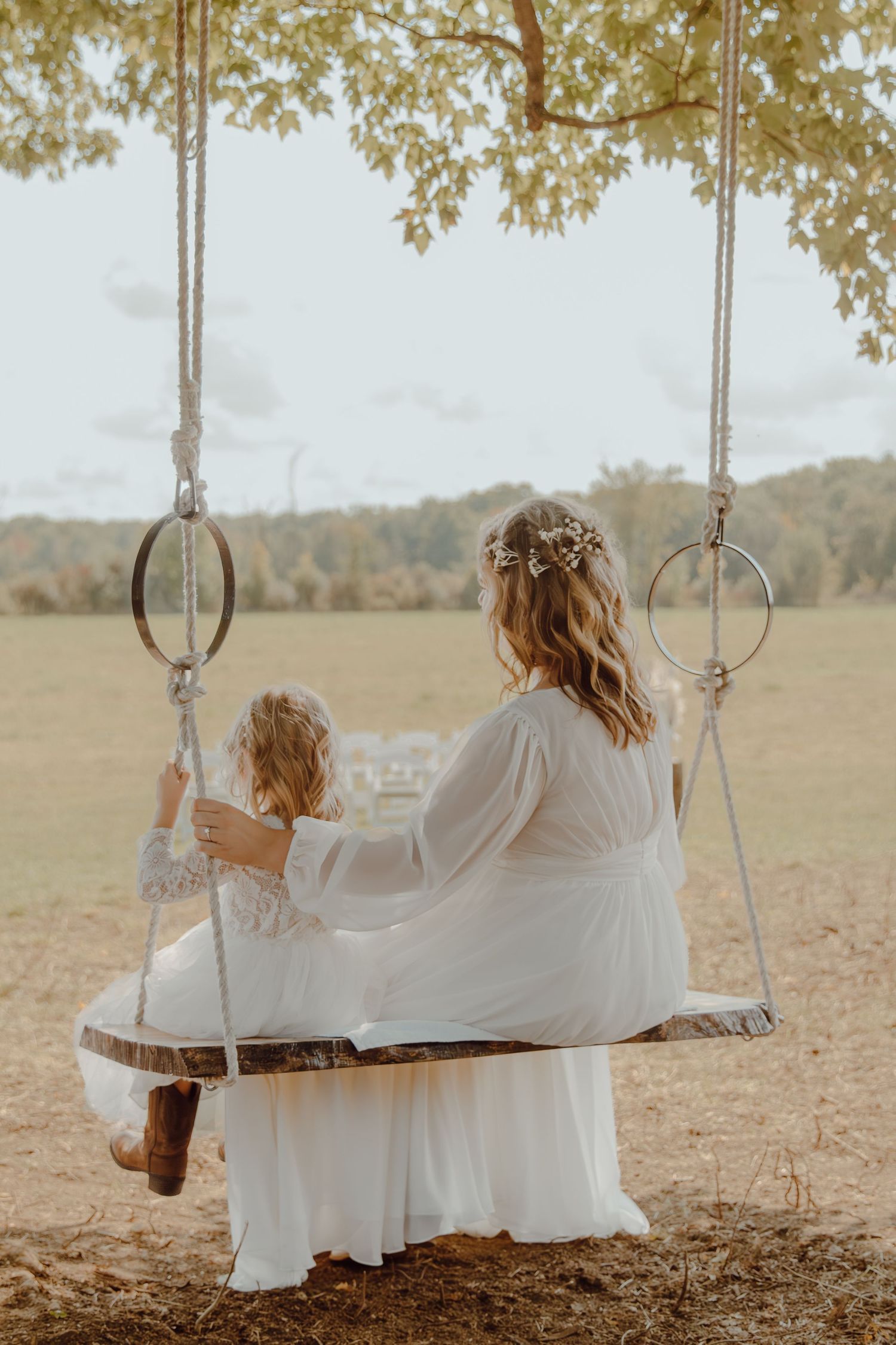 Two people in white dresses sit together on a rustic wooden swing in a peaceful outdoor setting.