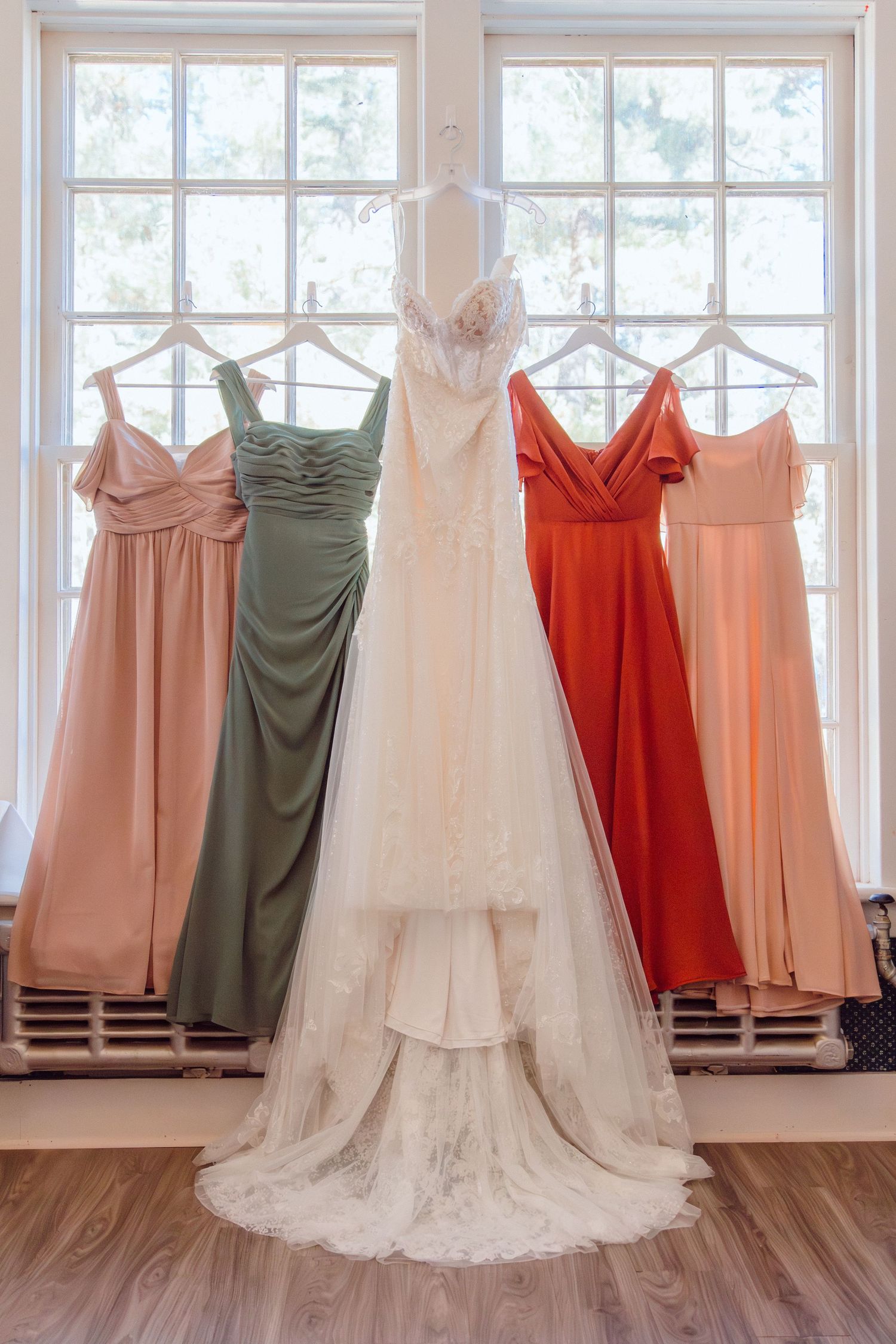 Wedding dress and colorful bridesmaid dresses hanging on wooden hangers against bright windows.