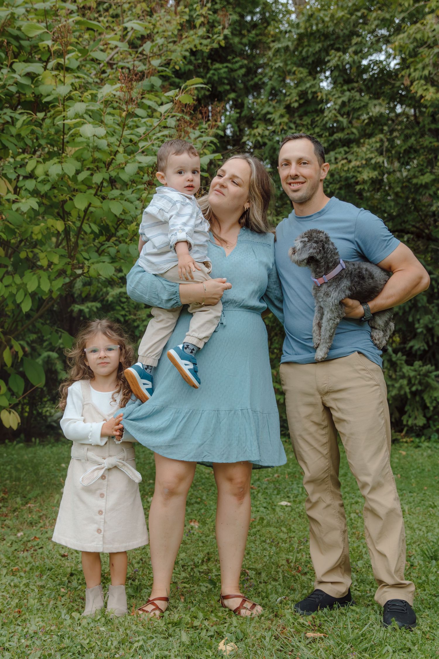 Family portrait in coordinated blue outfits with their dog in a park setting.