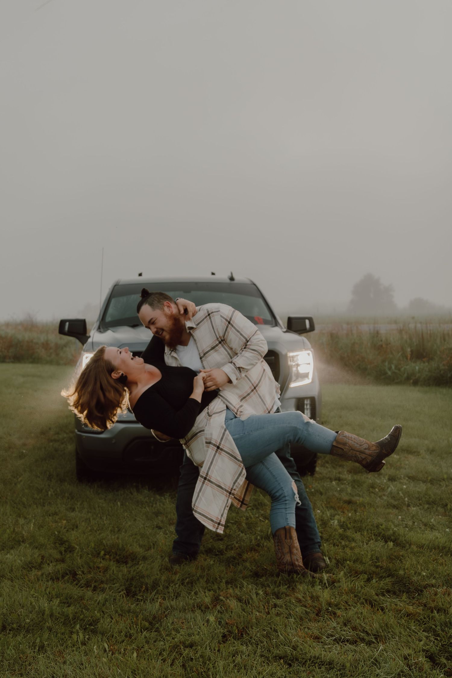 Couple dancing romantically in a foggy field near a pickup truck, woman lifted in mans arms, wearing casual country outfits.