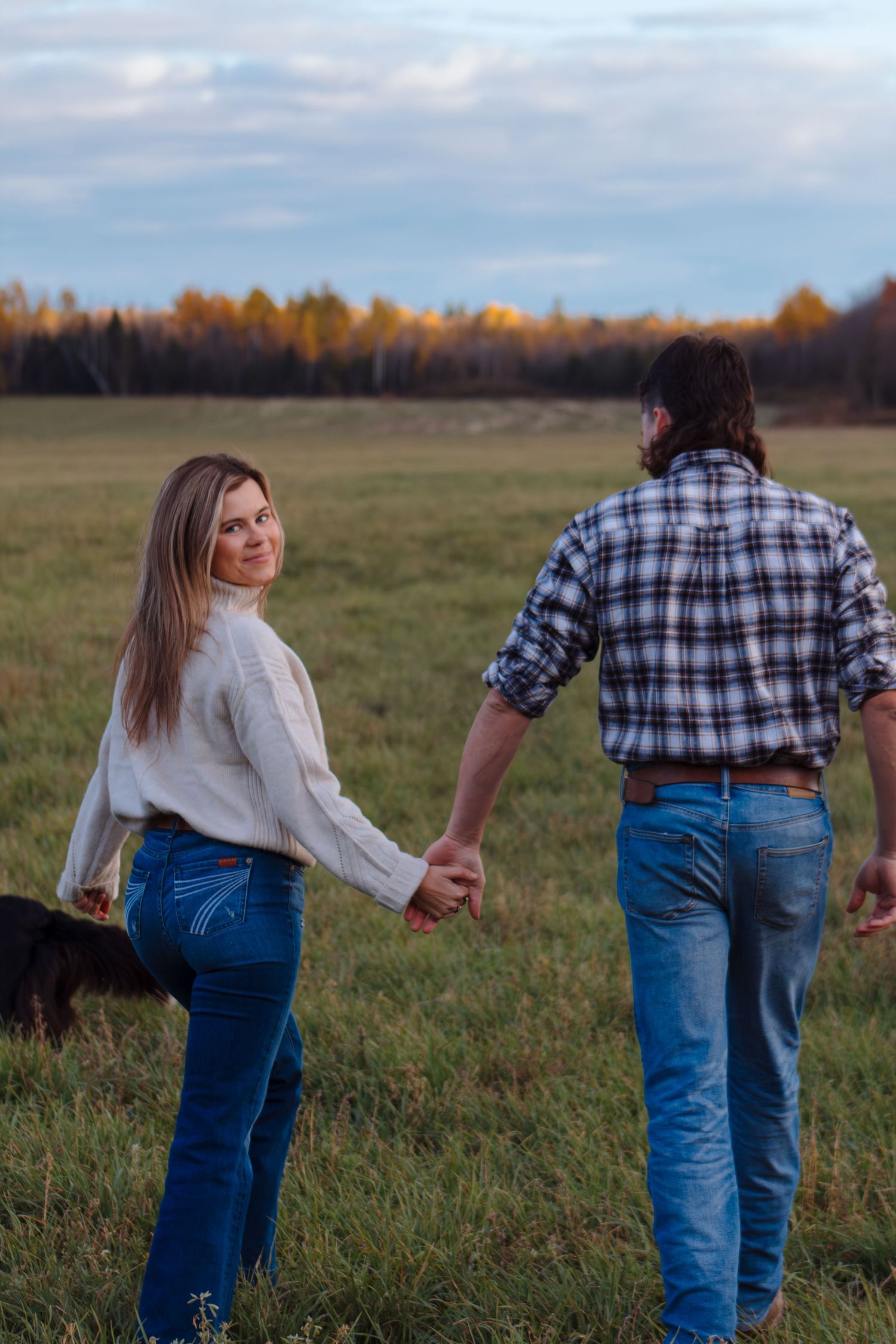 Two people hold hands while walking through a grassy field with fall foliage in the background.