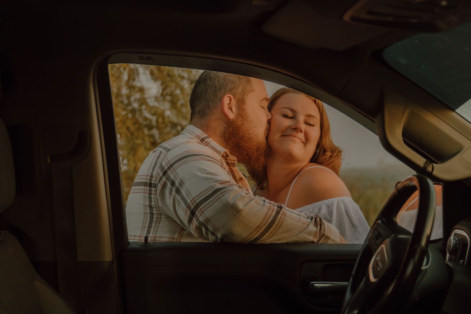 A couple shares a romantic embrace outside a car window, with the man kissing the woman's cheek at golden hour.