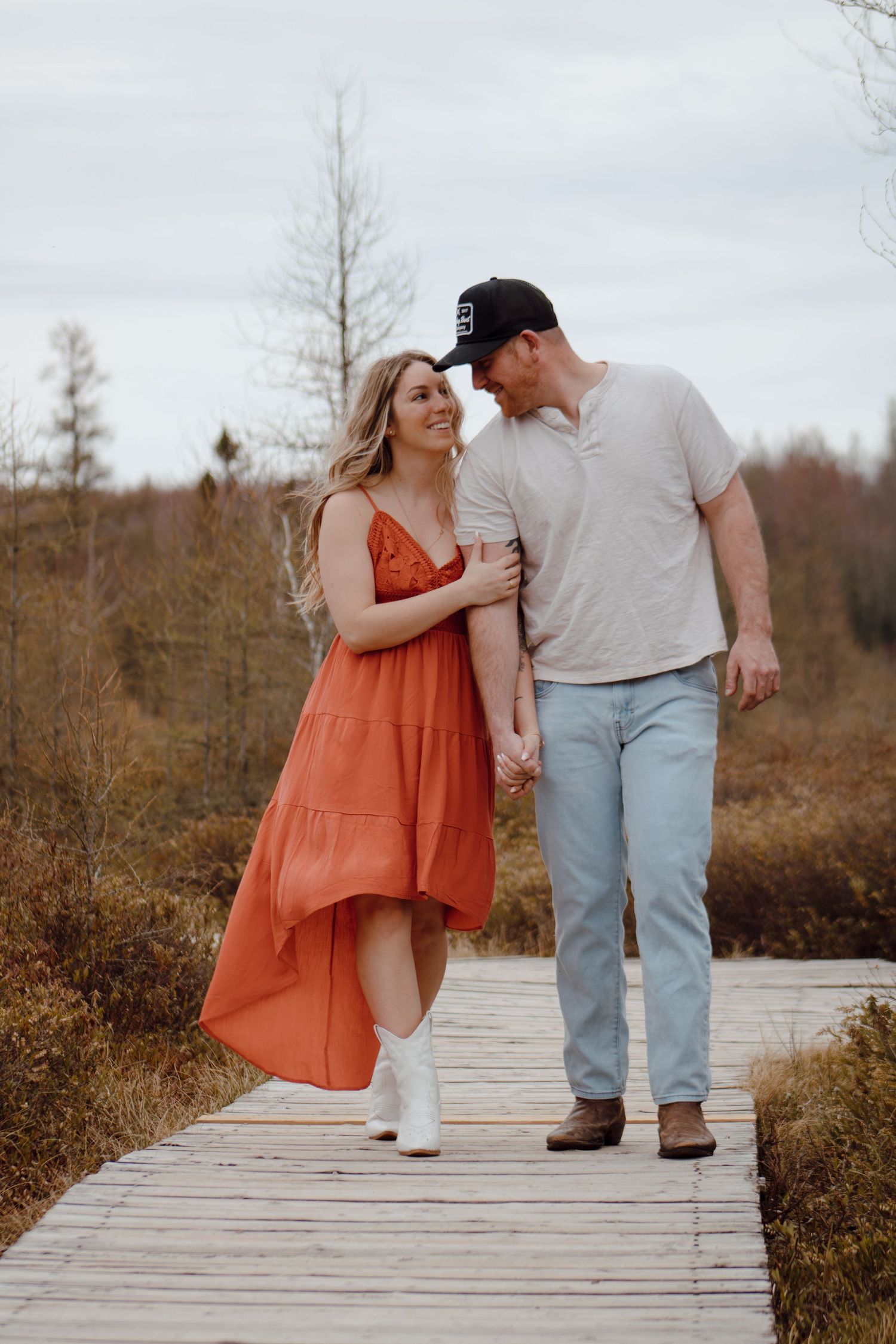 Couple walking hand-in-hand on a wooden boardwalk, woman in orange dress and white boots, man in jeans and cap.