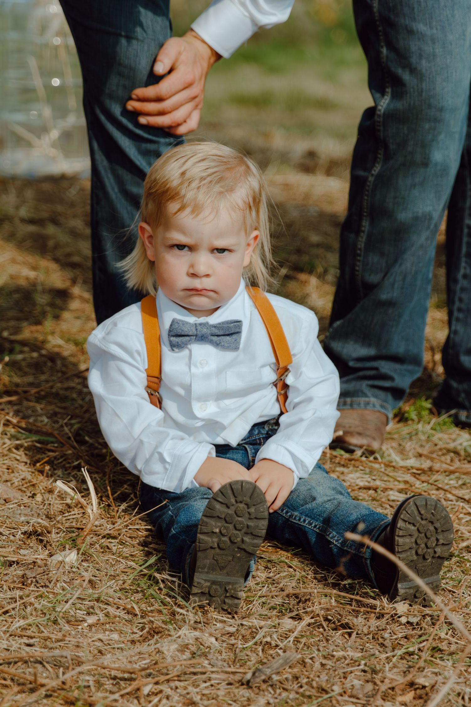 A young attendee in formal wear with suspenders and bowtie sits on the ground at an outdoor event.