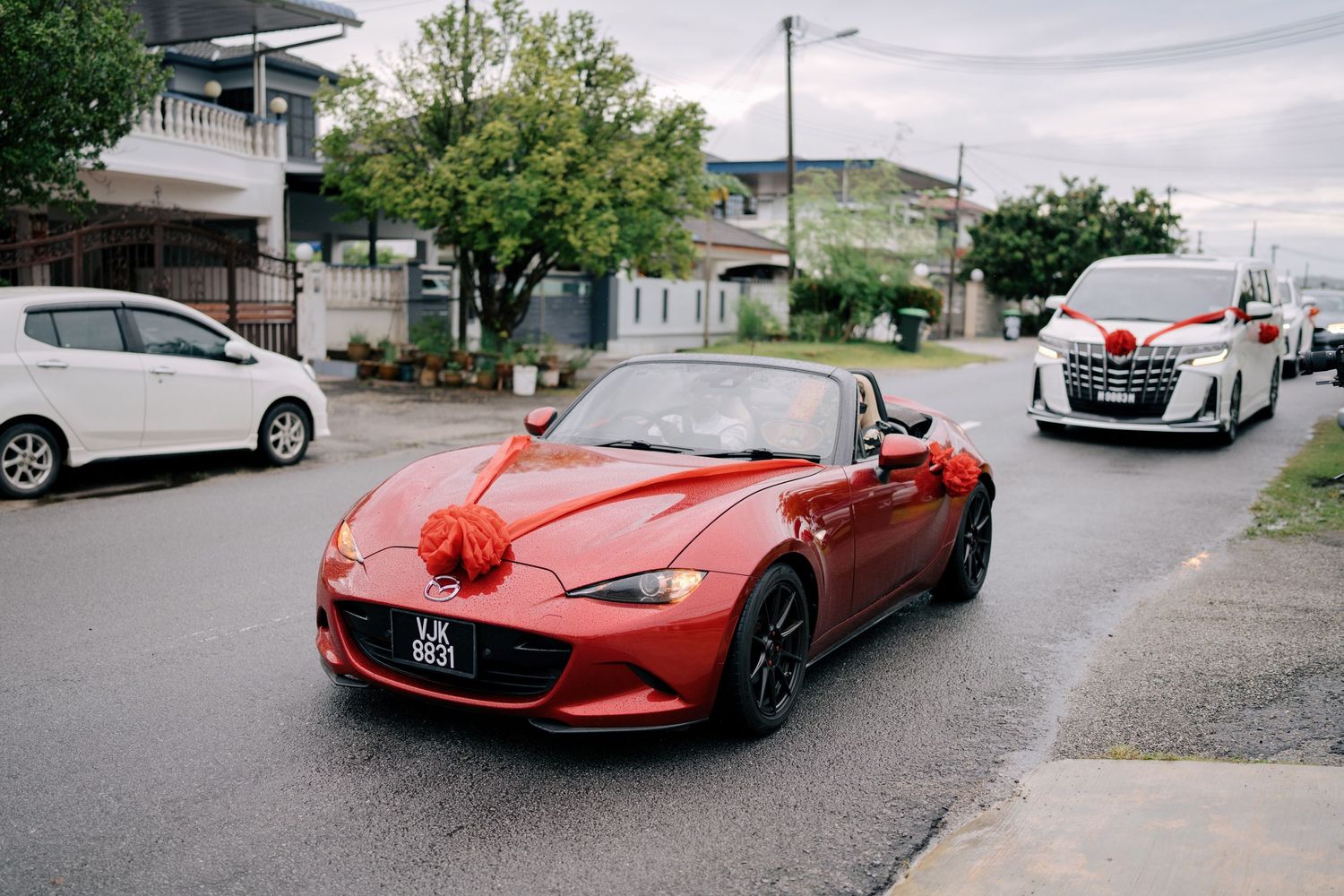 A red wedding car followed by a white luxury van driving through a residential neighborhood in Malaysia