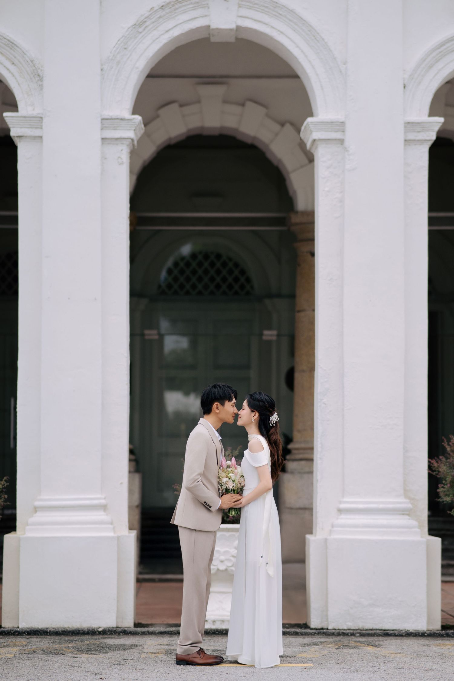 A couple shares a tender moment under white colonial arches of a historic Malaysian building.