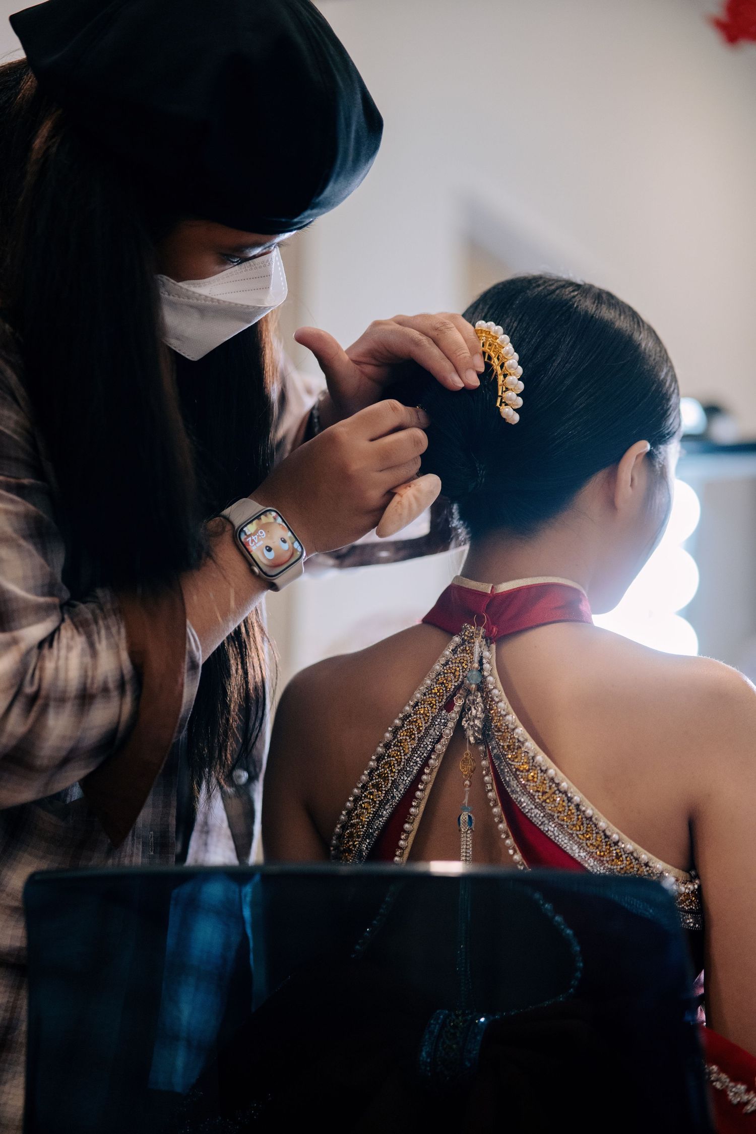 A hair stylist placing a gold and pearl comb into a bride's sleek bun, viewed from behind