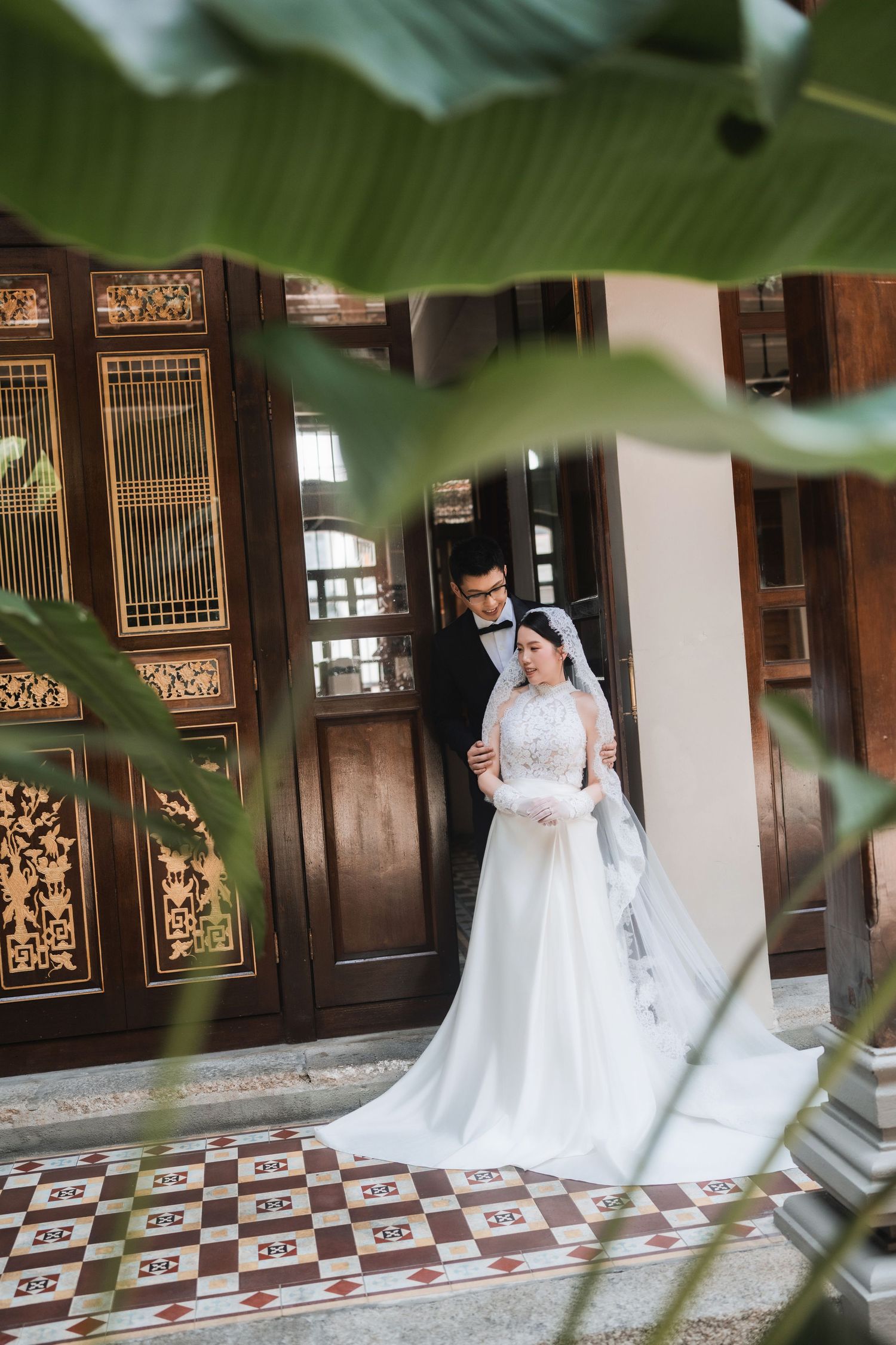 Wedding photography through tropical leaves showing couple in traditional Asian architectural setting.