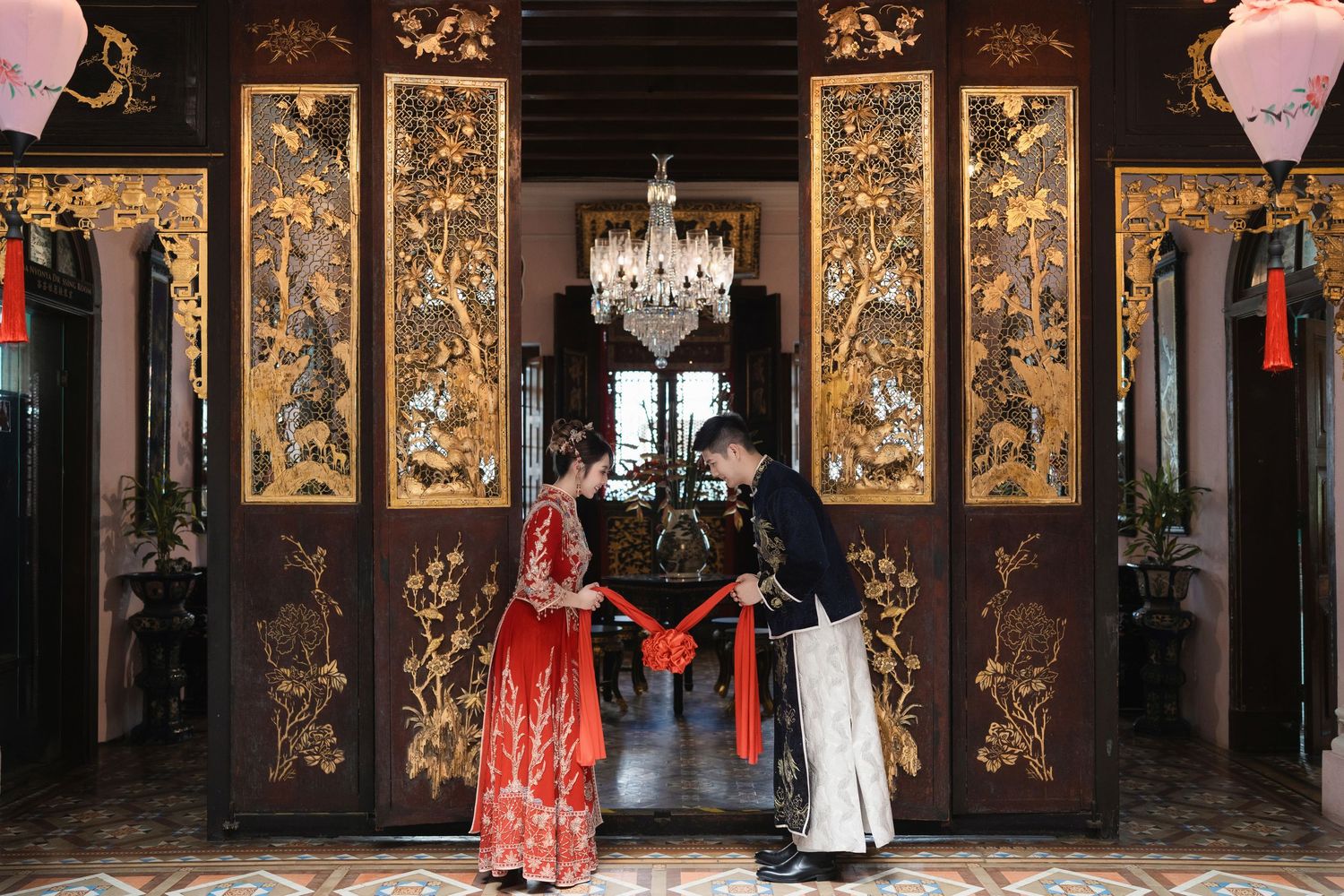 Couple performing a traditional wedding bow with a red silk ribbon between ornate gold-carved screens at Pinang Peranakan Mansion
