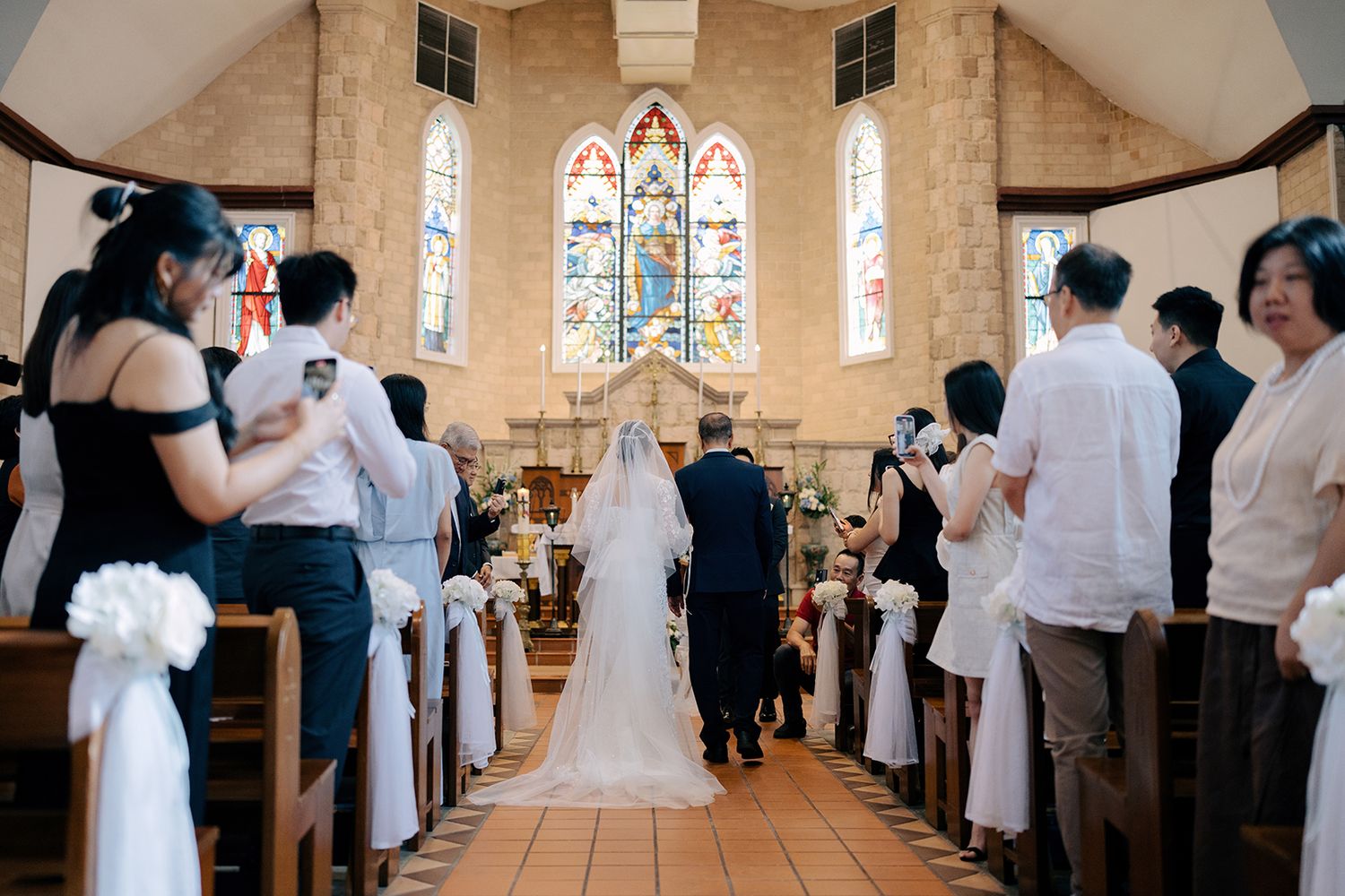View from the back of a church showing the bride in a long veil walking toward a brightly lit altar and stained glass