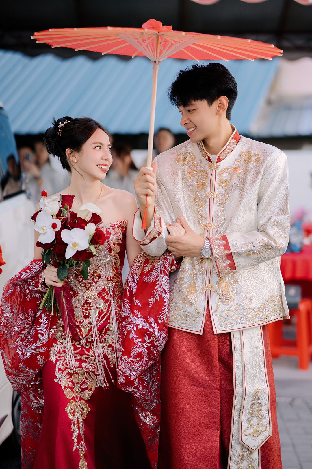 Couple in traditional red and gold Chinese wedding attire holding a decorative red parasol.